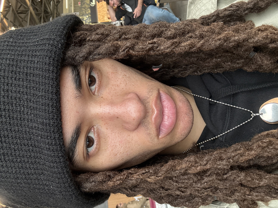 The image displays a close-up portrait of an individual situated within an indoor public area in Somma Lombardo, Italy. The central subject is a person with medium brown skin, marked by visible freckles on the nose and cheeks. They possess dark brown eyes, thick dark eyebrows, a prominent nose, and full, closed lips with a slight stubble visible on the chin and upper lip. The person wears a black knitted beanie covering their forehead and upper head, from beneath which numerous long, dark brown dreadlocks cascade around the face. They are dressed in a black hooded sweatshirt worn under a light-colored, possibly tan, collared jacket or coat. A silver-colored ball chain necklace is visible, featuring a pendant composed of an oval silver disc layered over a larger, oval-shaped element, possibly made of wood or copper.
The background, partially out of focus, reveals an expansive indoor space with a high ceiling characterized by visible metal support structures or trusses. Blurred figures of other individuals are discernible in the mid-ground, including a man with a beard and glasses partially visible on the right side of the frame, appearing to be seated. A fragmented commercial sign displaying the letter "V" and subsequent letters such as "ene" or "enice" is visible in the upper right background. The overall environment, with its structural elements and presence of multiple people and commercial signage, is consistent with an airport terminal, specifically Milan Malpensa Airport, which is located in Somma Lombardo.