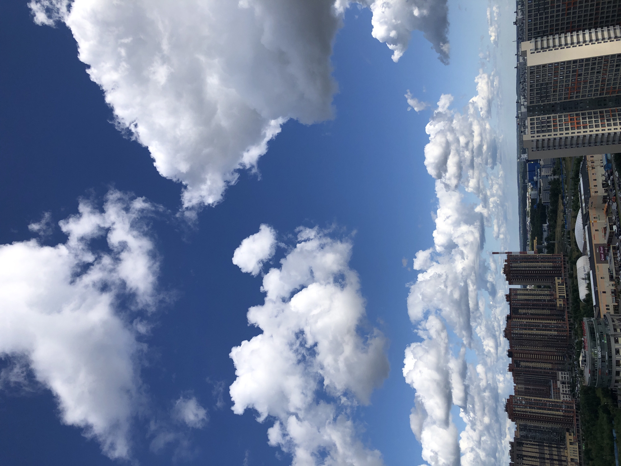 The image depicts a wide-angle view of a city skyline under a bright blue sky filled with fluffy white clouds. In the foreground, a cluster of tall, modern residential buildings with a beige and orange facade dominates the scene. These buildings are densely packed, with numerous windows visible. To the left, another row of similarly tall buildings, primarily in shades of brown and red, is also visible.
The middle ground shows a mix of lower-rise buildings, some appearing to be commercial or industrial, with a large white domed structure and a red and white striped smokestack. A sprawling green area with trees separates these structures from the residential towers. In the background, a hazy cityscape extends to the horizon, suggesting a vast urban area. The sky is a vibrant blue, with a significant portion occupied by large, cumulus clouds, some casting faint shadows. The overall impression is one of a contemporary urban environment on a clear, partly cloudy day.