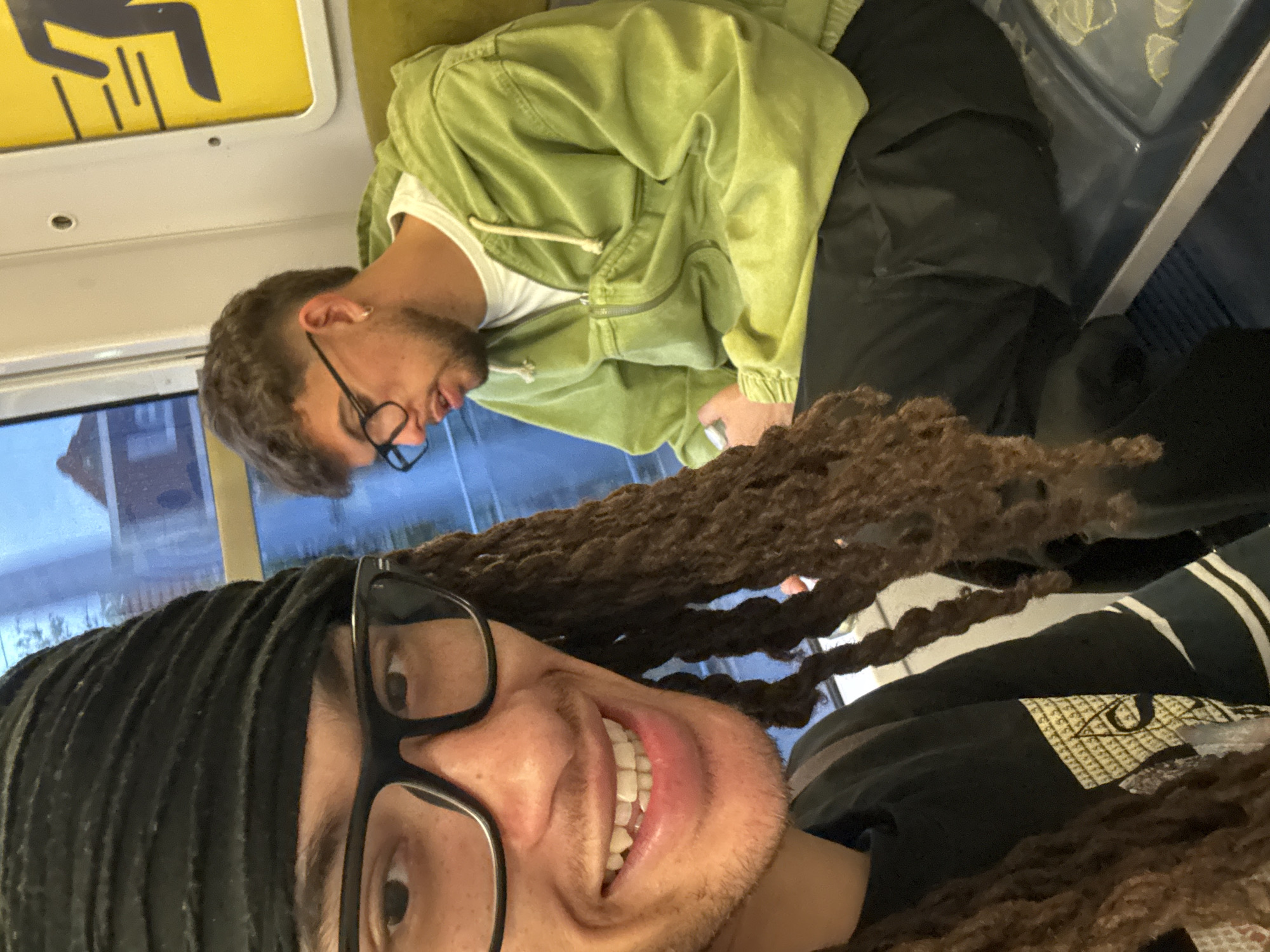 The image captures a selfie taken inside what appears to be a train or metro carriage in Orsay, France, featuring two men.
In the foreground, the person taking the selfie smiles broadly, showing teeth. They wear thick-framed black glasses and have dark dreadlocks or braids partially covered by a black durag or headwrap. They also have some facial hair. Their dark t-shirt features a distinctive graphic design: an eye within a pyramid or triangle, set against a checkered pattern, along with horizontal stripes on the upper part of the shirt.
In the background, another man is seated on a green fabric bench, looking downwards or to the side, with a slight smile. He has short dark hair, a beard, and wears thin-framed black glasses and a small earring in his left ear. He is dressed in a light olive-green hooded jacket over a white t-shirt and dark trousers.
The setting is a public transport interior, indicated by the visible window on the left, which reveals a blurred view of residential buildings or an urban landscape outside. The ceiling of the carriage is visible, along with a yellow sign featuring a black pictogram, likely an accessibility symbol or indication of a priority seat. The seats appear to be a mix of green and blue/dark upholstery. The overall lighting suggests it could be daytime, possibly overcast, or early evening. The foreground person's jovial expression contrasts with the background person's more relaxed and neutral demeanor.