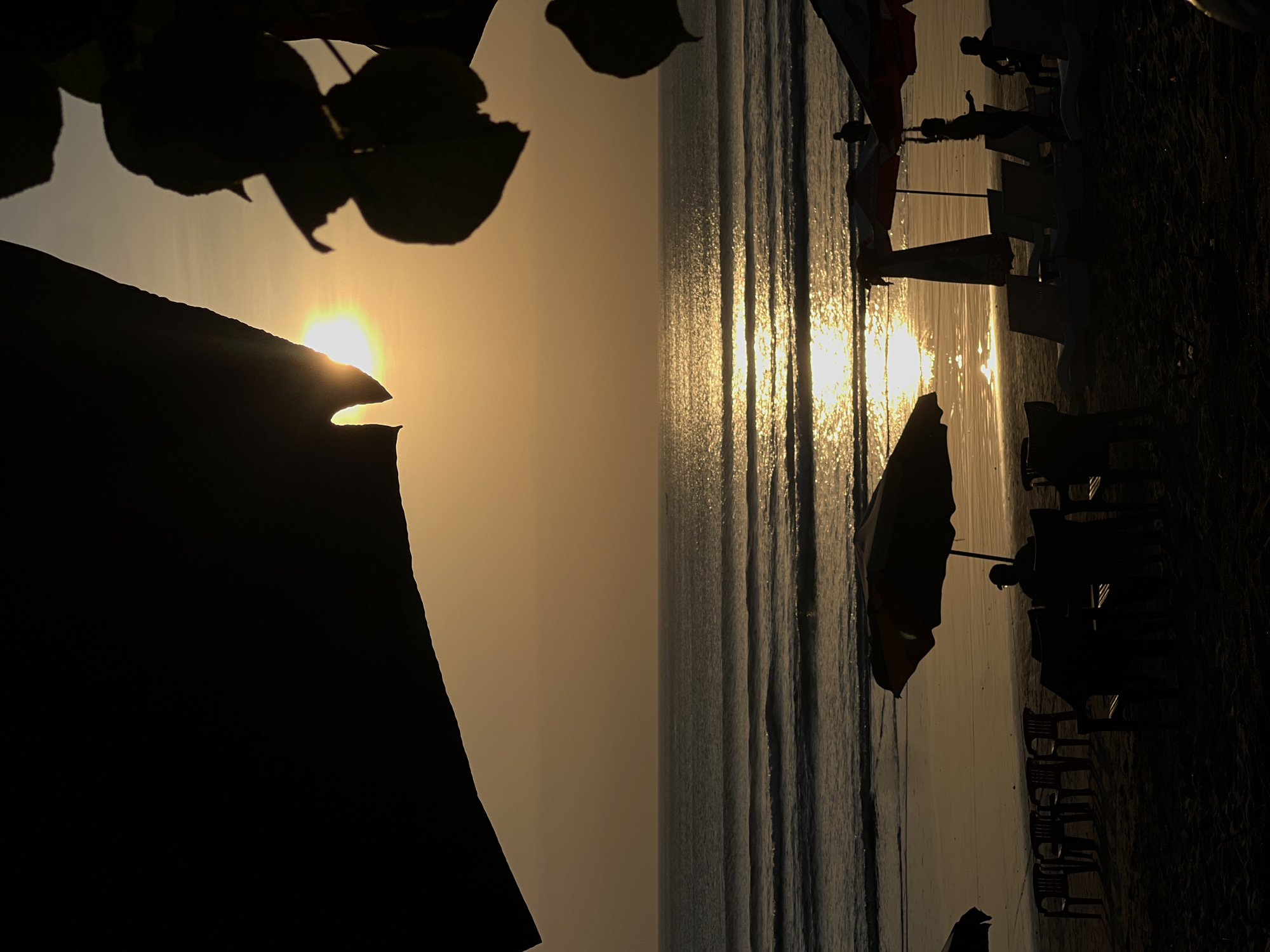 The image depicts a serene sunset scene on Kuta Beach, Indonesia. The sun, a bright orb of light, is partially obscured by dark silhouettes of foliage and a large, curved object in the foreground, casting a warm, golden glow across the sky and water. Gentle waves roll onto the sandy shore, reflecting the sunlight in shimmering patterns. In the mid-ground, several beach umbrellas and lounge chairs are arranged on the sand, some occupied by silhouetted figures. To the left, a row of chairs is set up, facing the ocean. To the right, more lounge chairs and umbrellas are visible, with a few people scattered around. The overall atmosphere is tranquil, capturing the essence of a peaceful evening at this popular Indonesian beach.