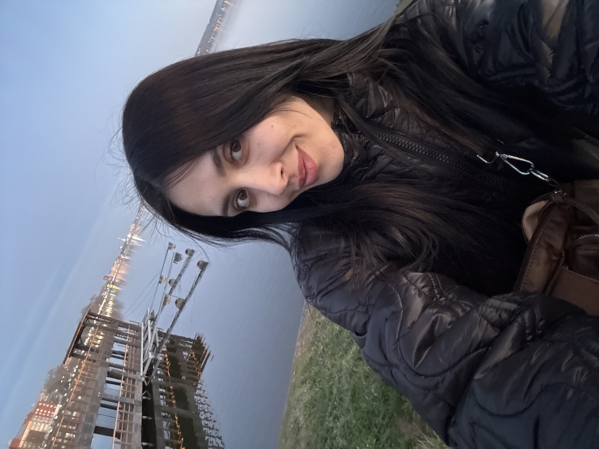 A woman is standing outside, possibly near the waterfront. She is wearing a black, puffy jacket, and has long, dark hair. In the background, there is a body of water with what appears to be a city skyline or construction site visible across it. To the left of the woman, there is a pier and dock, with multiple bright lights turned on. The grass is also visible in the image. Given the request to include Votkinsk, Russia, the woman is likely located near the Votkinsk Reservoir or another prominent waterfront in the city.