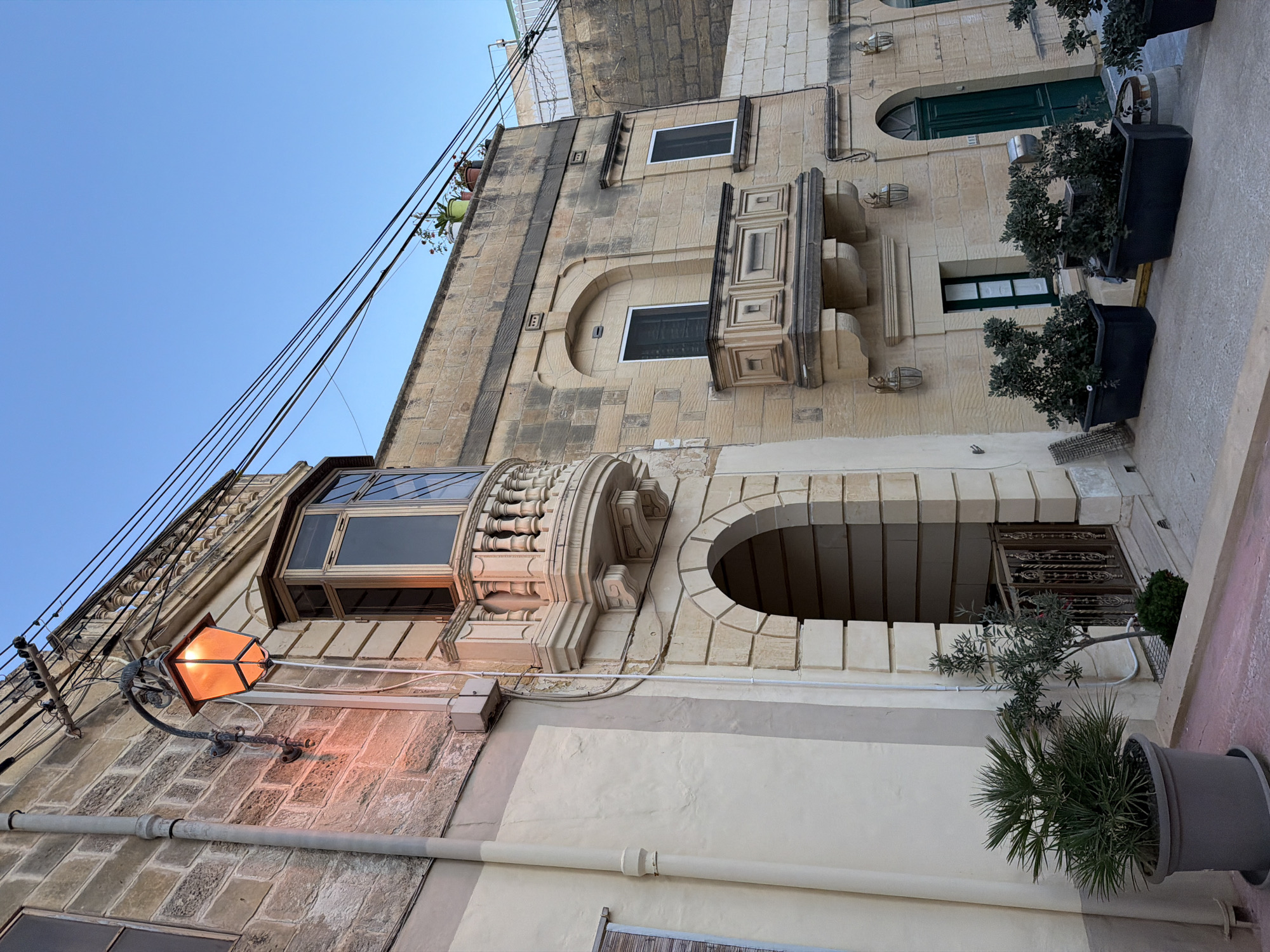 The image depicts a street scene in Fontana, Malta, featuring traditional Maltese architecture. A stone building with a pale yellow facade dominates the foreground. A prominent feature is an arched entryway on the lower level, revealing a decorative metal gate. Above this, a bay window with a stone balustrade juts out from the building. An ornate, orange-lit street lamp is mounted on the wall to the left of the bay window, casting a warm glow. The building's facade is constructed from weathered, light-colored stone blocks, characteristic of Maltese construction. A smaller, darker stone building is visible to the right, with a green door and windows. Potted plants are strategically placed along the sidewalk in front of both buildings, adding greenery to the scene. Overhead, a network of electrical wires crisscrosses the clear blue sky. The overall impression is one of a historic and charming Mediterranean street.