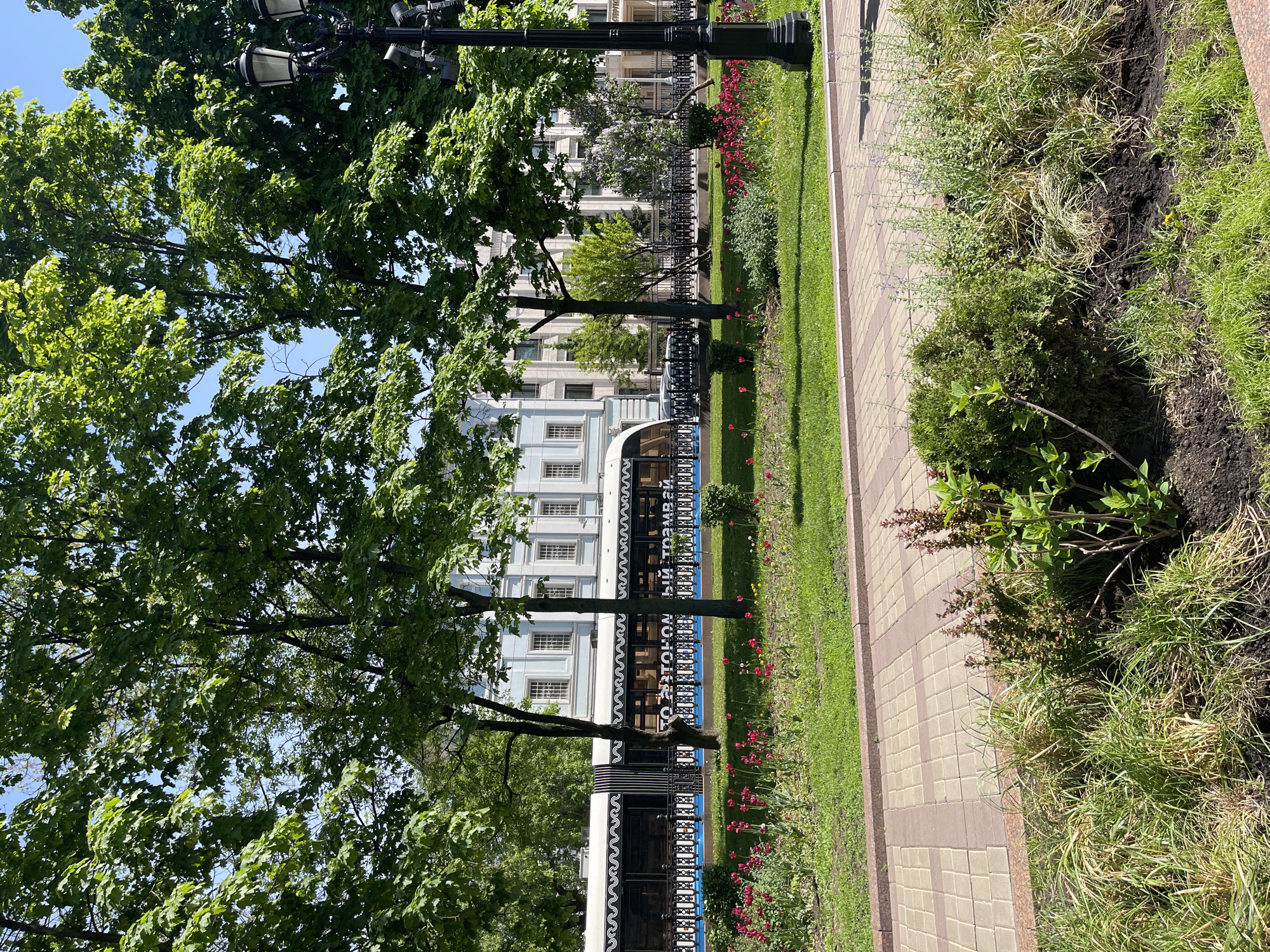 The image shows a sidewalk lined with landscaping and trees, with a building visible in the background. The sidewalk is paved with interlocking bricks. Landscaping includes grass, flowers, and small bushes. Trees are mature and leafy. A street lamp is visible. The building has a light blue façade with white trim and many windows. A tram is partially visible in front of the building, with text in what appears to be Russian.