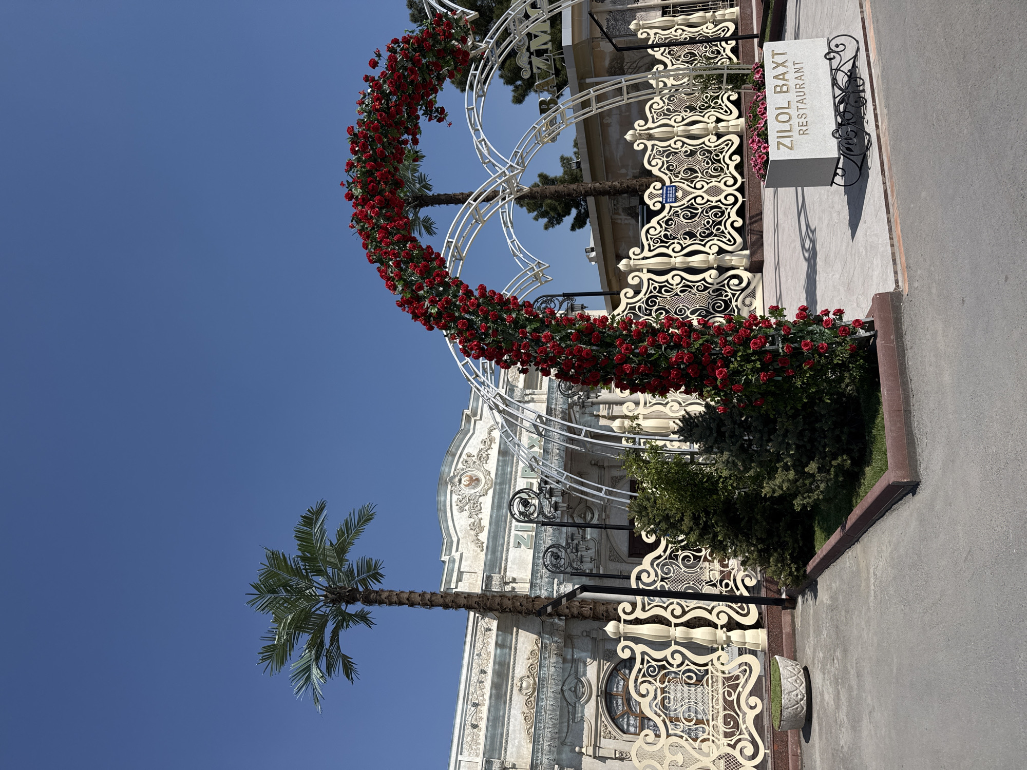The image depicts the entrance to the "ZILOL BAXT Restaurant" in Samarkand City, Uzbekistan. A white metal archway adorned with red roses frames the entrance. A palm tree stands to the left of the archway. The restaurant building features ornate white architectural details. A sign bearing the restaurant's name stands to the right. The sky is clear and blue. The overall scene suggests an upscale or decorative establishment.