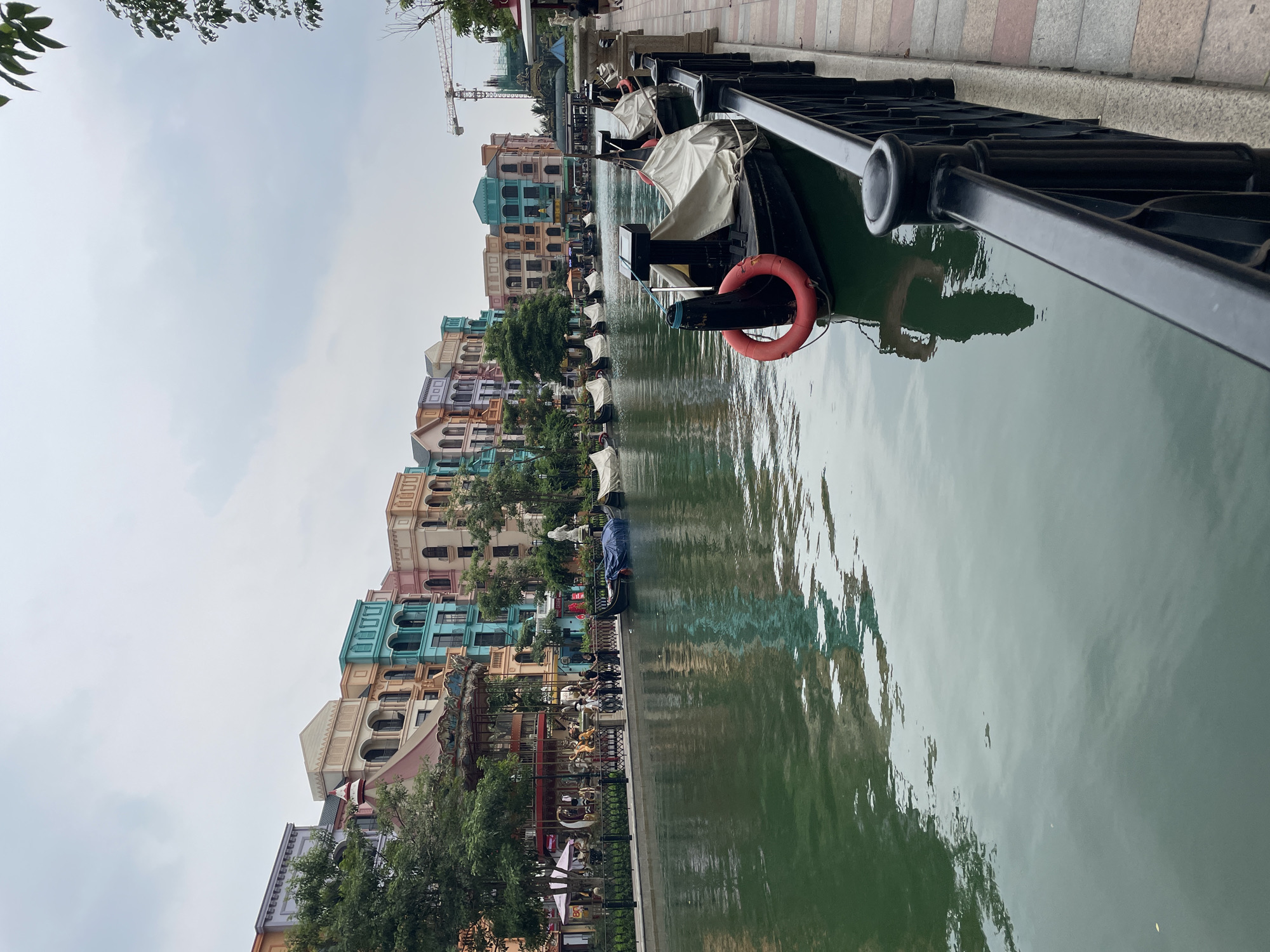 The image depicts a canal lined with colorful buildings in the city of Nghĩa Trụ Commune, Vietnam. The architecture appears to be inspired by European styles, with pastel-colored facades and ornate details. Several gondolas, covered with white tarps, are moored along the canal, suggesting a tourist attraction or a themed area. One gondola in the foreground is uncovered, showing a red life preserver. People are visible on the walkway beside the canal, some standing near a carousel. The water in the canal is a deep green, and its surface reflects the buildings and sky. A black metal railing runs along the edge of the walkway. In the background, a construction crane is visible, indicating ongoing development. The sky is overcast, with a muted, diffused light. Trees and greenery are interspersed between the buildings.