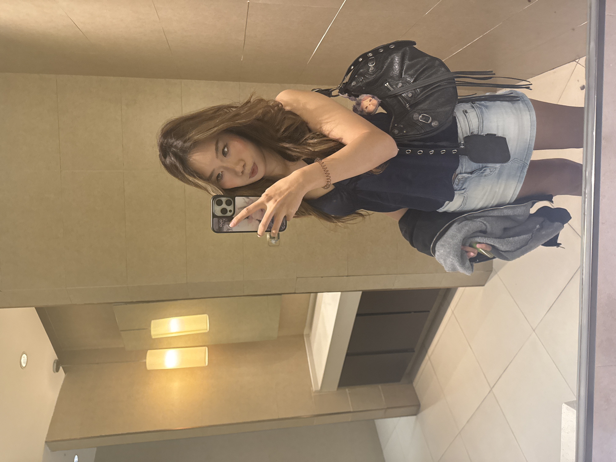 A woman with brown hair takes a selfie in a bathroom mirror. She is wearing a dark top, denim skirt, and tights. A black studded purse hangs over her shoulder. The bathroom has beige tiled walls, rectangular lights above the sink, and a white countertop with a modern sink. The floor is tiled in a lighter color. The location is likely not Yuzhong District, China, as the style and architecture of the bathroom do not suggest a location in China.