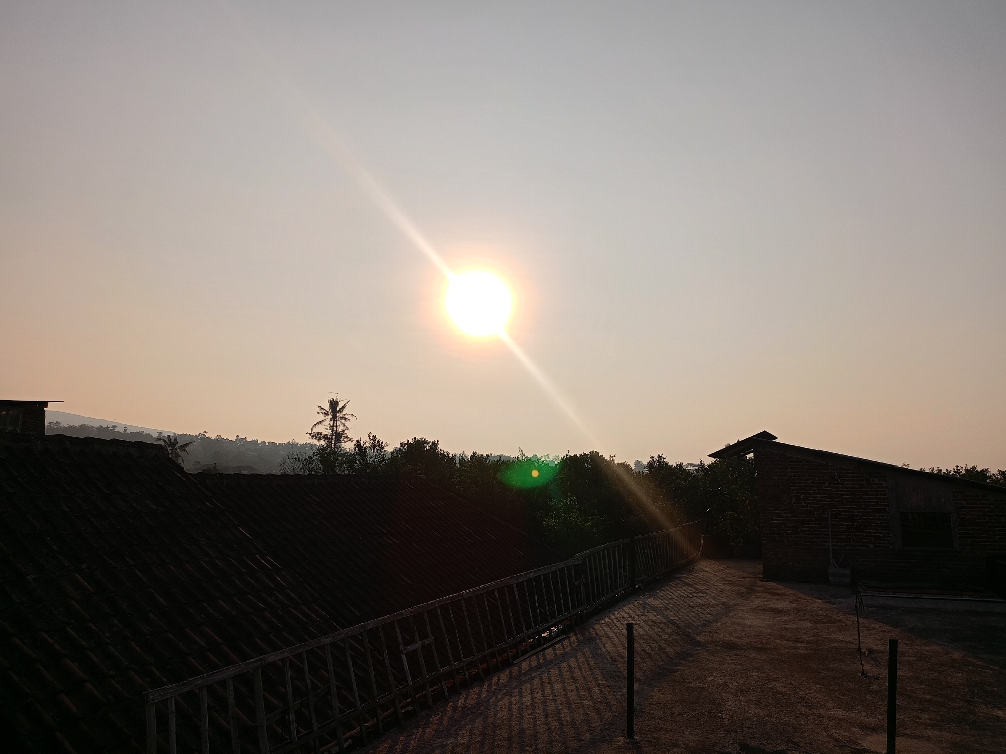 The image depicts a sunset scene from a rooftop in Batu, Indonesia. The sun is a bright, almost blinding disc in the pale sky, casting long, sharp rays downwards. A faint green lens flare is visible. In the foreground, a tiled roof of a building occupies the left half of the frame. A wooden railing runs along the edge of the rooftop, casting a grid of shadows onto the concrete surface. To the right, a brick building with an open roof structure stands partially silhouetted. The background is filled with a dense line of trees and vegetation, with rolling hills faintly visible in the distance, shrouded in mist. The overall impression is one of a tranquil, rural evening.