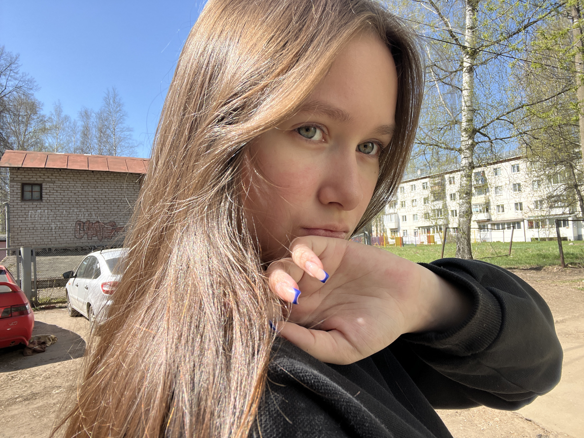 A young woman with long, light brown hair is the subject of a medium shot, slightly angled to the left. She is wearing a black sweatshirt and has blue nail polish on a manicure. Her hand is lightly touching her chin. The background includes a weathered brick building with a rusty brown roof, parked cars (red and white), and an apartment building with white walls and windows. Trees with budding leaves fill the remaining spaces. The photograph was taken in a public space during the day in the city of Chaikovsky, Russia. The sunlight suggests the time is around midday.