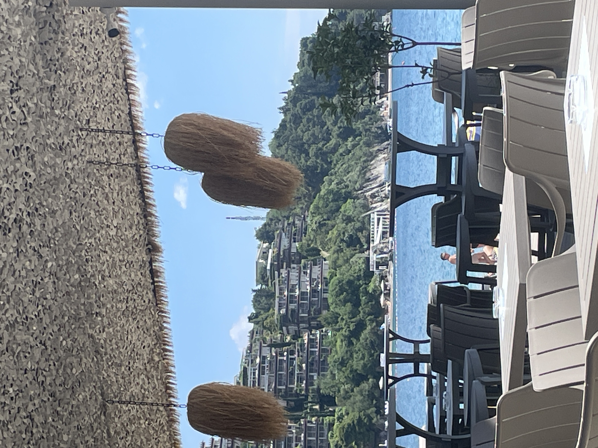 The image shows an outdoor seating area, likely a restaurant or cafe, overlooking the Adriatic Sea in Budva, Montenegro. The scene features tables and chairs under a thatched roof decorated with hanging straw ornaments. In the background, a hill is visible with several buildings, indicating a developed area along the coast. A person is discernible by the sea. The setting suggests a tourist-oriented location.