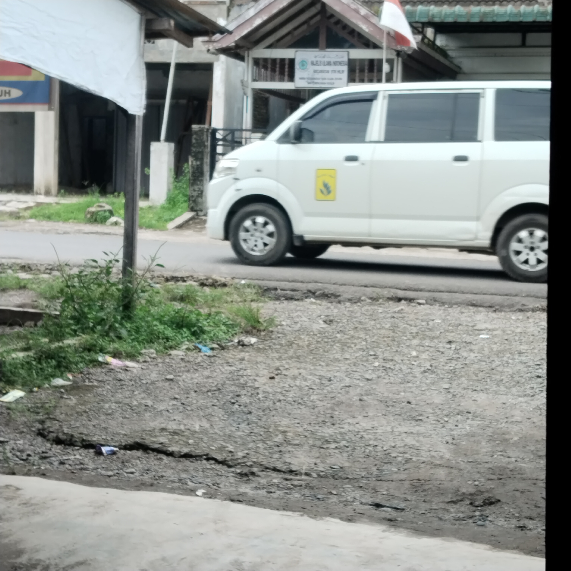 A white van is parked in front of a building in Talun Kenas, Indonesia. The building has a dark-colored roof with green trim. A small Indonesian flag is mounted on a pole in front of the building. The van has a yellow emblem on its side. The ground in front of the building is covered with gravel and some debris, including a cracked pavement area. There is also some vegetation growing near the building and on the side of the road. The scene appears to be a public space in a town or village.