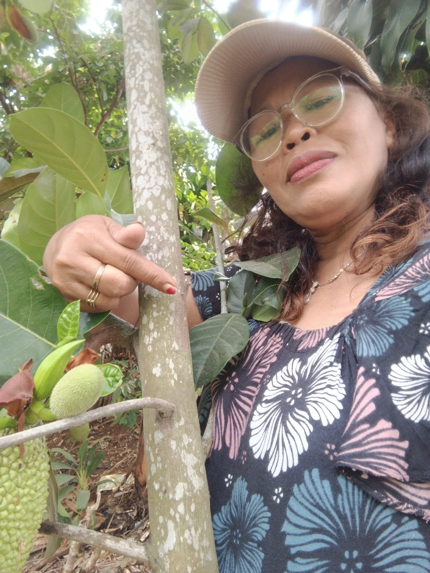 A middle-aged Indonesian woman with curly brown hair, wearing a tan baseball cap and glasses, poses for a selfie in an outdoor environment, likely a garden or orchard, in Warungkiara, Indonesia. Her hand, adorned with two gold rings and red nail polish, points towards a jackfruit tree. The woman is wearing a dark top with a floral print of white, pink, and teal hibiscus flowers. In the background, lush green leaves and branches of the jackfruit tree are visible, along with a developing jackfruit. The sunlight filters through the leaves, creating a bright, natural lighting effect.