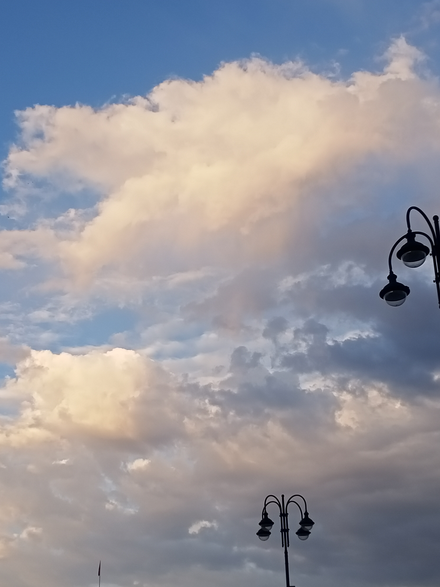 A low-angle shot captures a cloudy sky in Bishkek, Kyrgyzstan. The clouds are varied, with some appearing bright and illuminated by sunlight, while others are darker and more ominous. In the foreground, two street lamps are visible. The upper lamp is a single pole with two lights, while the lower lamp is a taller pole with four lights arranged in a cluster. The sky dominates the frame, suggesting a dramatic weather scene or sunset.