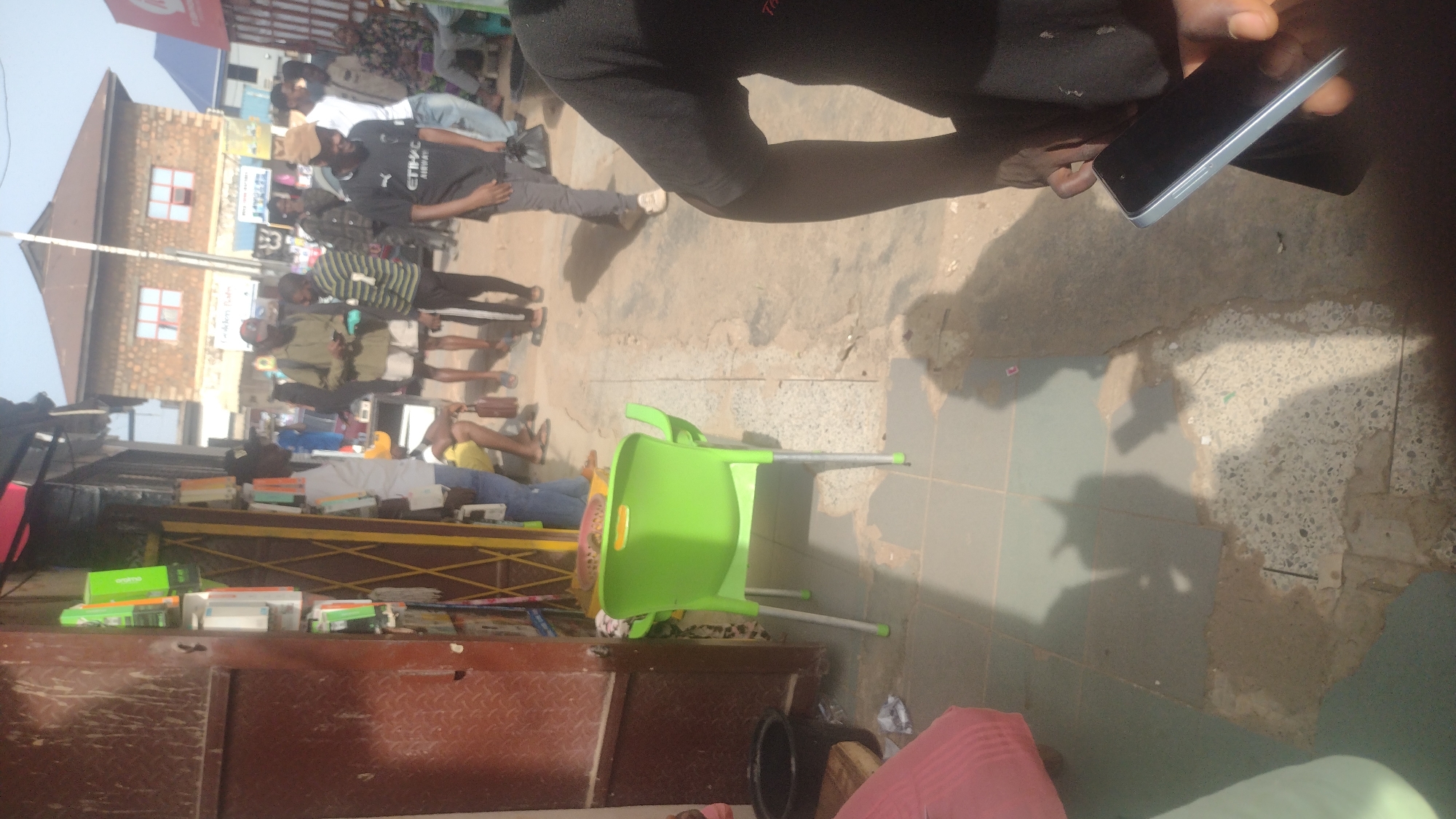 The image appears to be taken in Jos, Nigeria, showing a street scene. A group of people is visible further down the street. Several people are standing, some wearing masks. A light green plastic chair lies on its side. A table is laden with books and boxes of unknown content. A person is holding a cell phone. The ground is uneven and mostly covered with sand. A building stands in the distance.