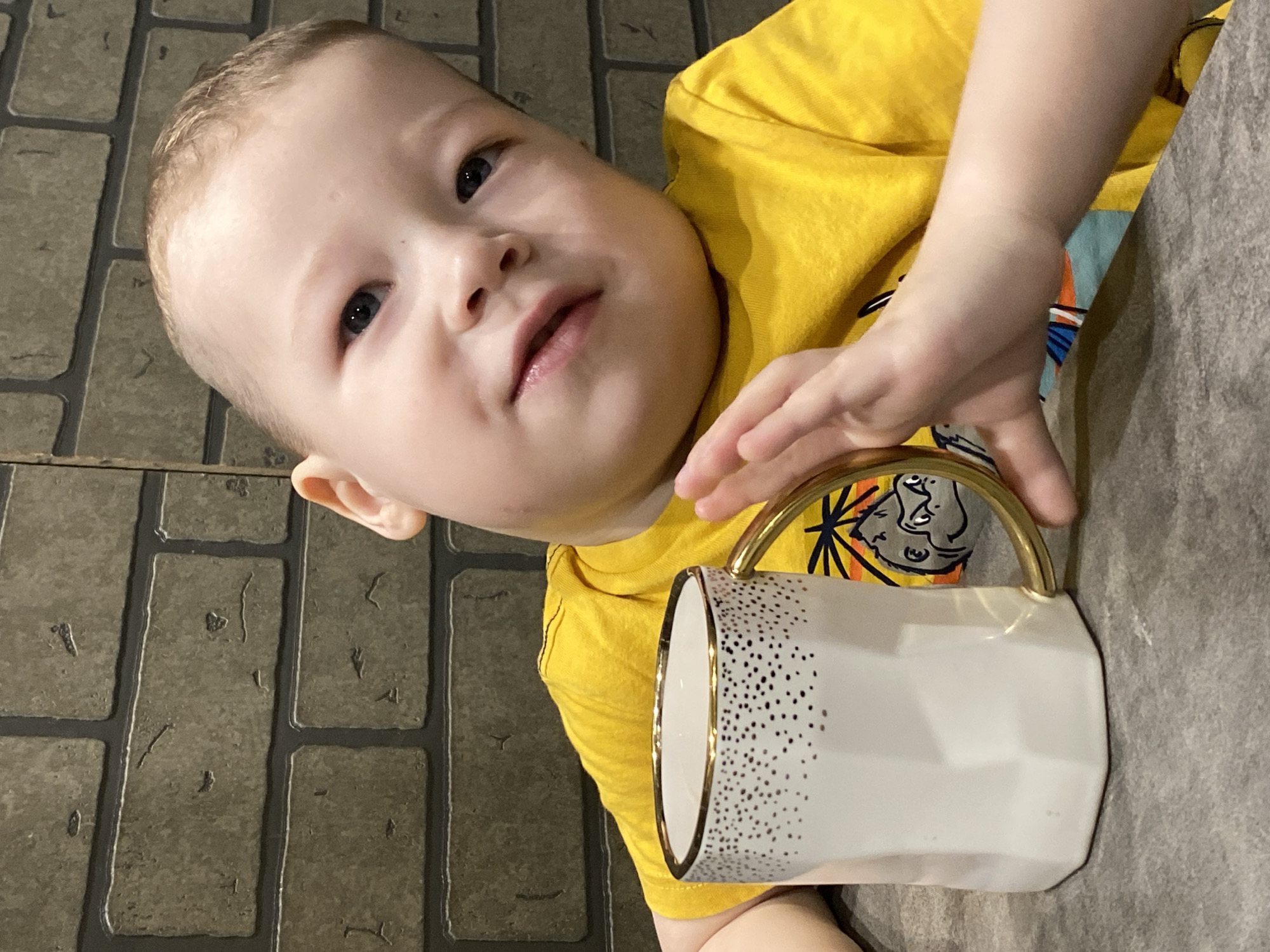 A young child, wearing a yellow t-shirt with a cartoon graphic, is positioned at the bottom of the frame. The child's face is turned upwards and slightly to the left. Their right hand is resting on the gold handle of a white mug decorated with black dots, which is located at the right center of the image. The mug is on a grey surface. The background features a brick-like pattern. The location is unknown and cannot be identified as Sochi, Russia from this image.