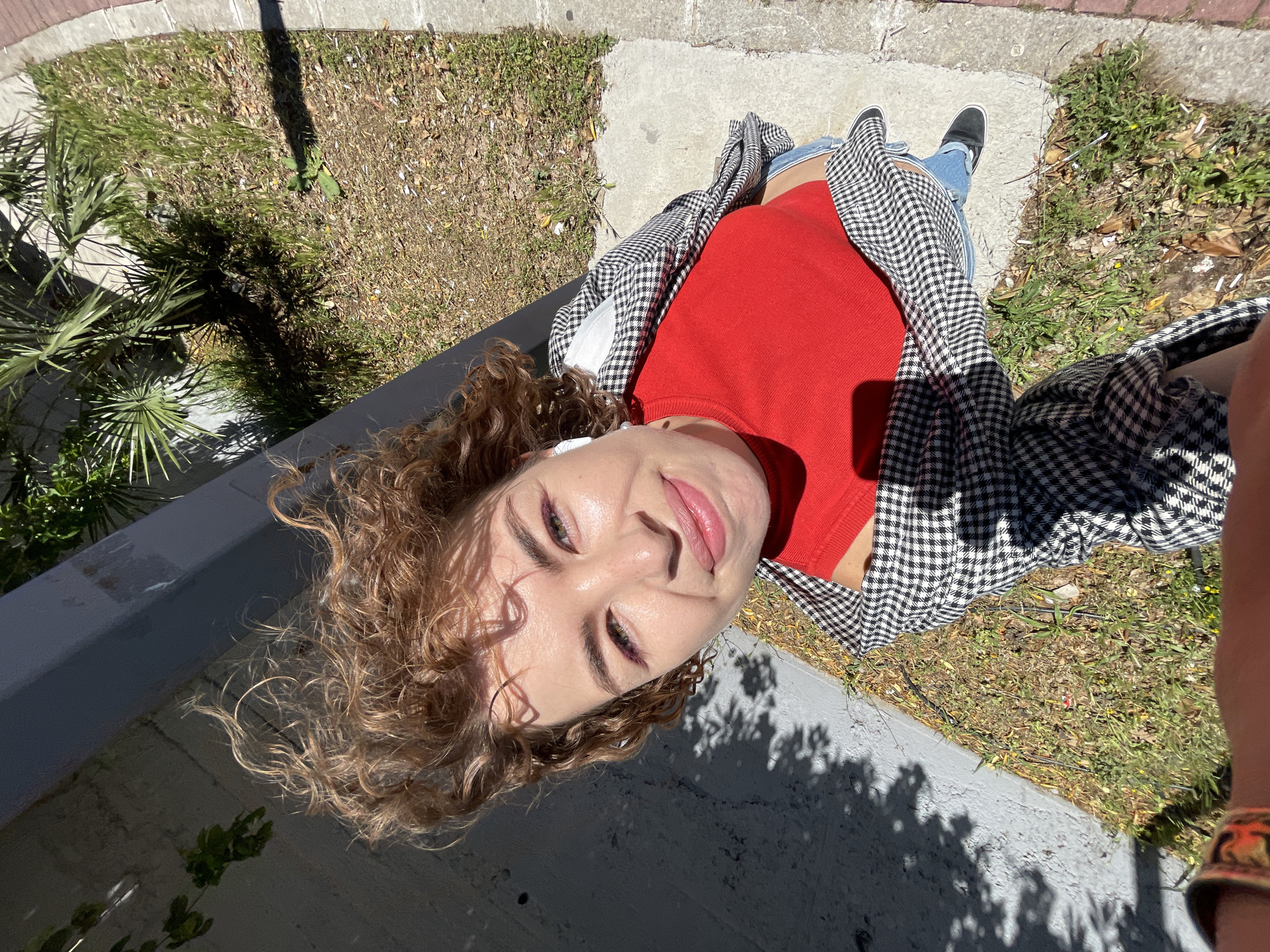 A high-angle shot shows a woman with curly hair, wearing a red shirt and a black and white checkered jacket. She is standing on a concrete surface, likely a sidewalk or ledge. Visible behind her are patches of grass, some concrete blocks, and foliage, possibly trees or shrubs. The lighting suggests it's a sunny day, with shadows cast across the concrete and greenery. In the background, there's a glimpse of what appears to be urban vegetation. There's no explicit indication of the location being Rafailovići, Montenegro.
