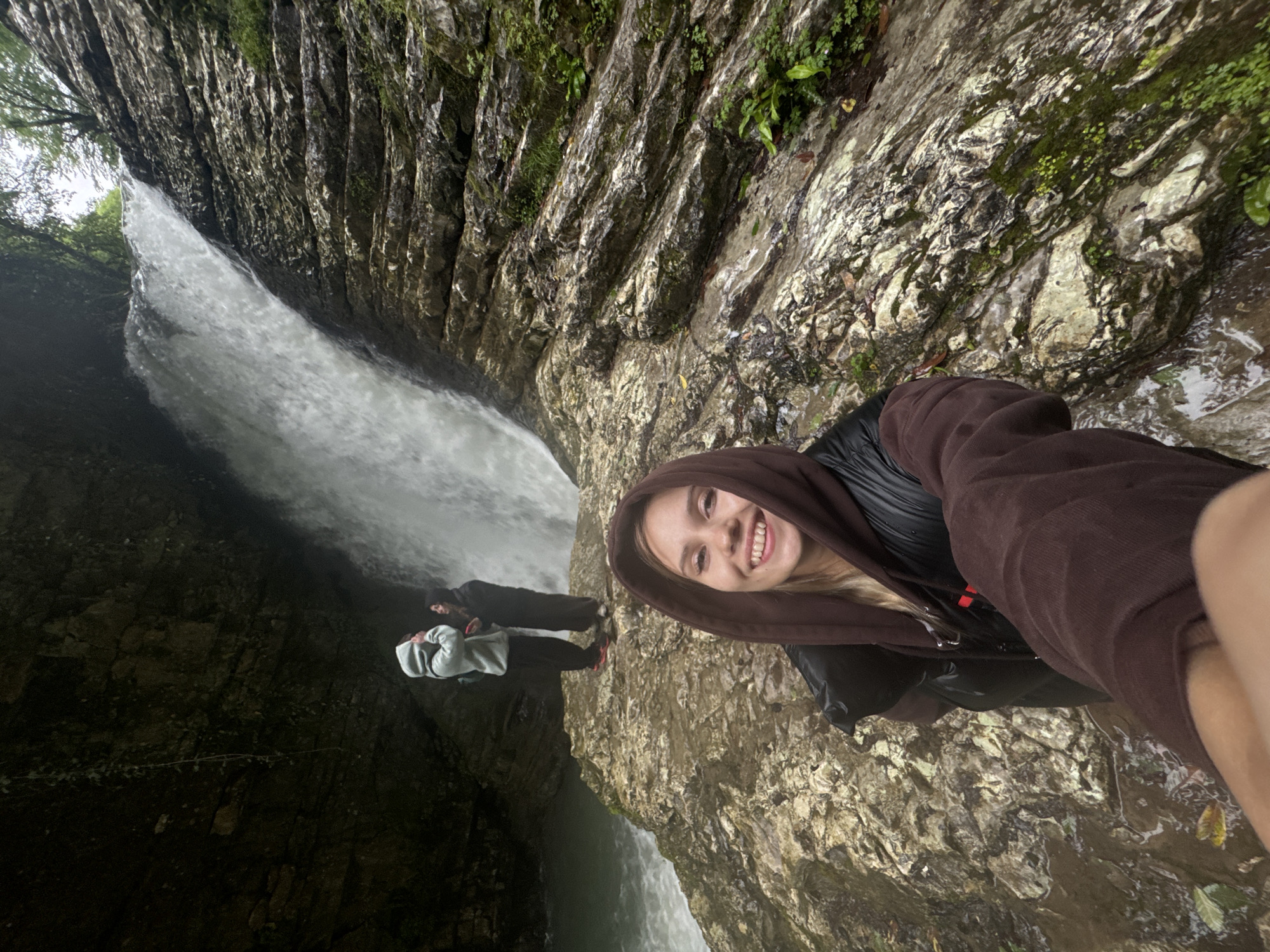 A woman in a brown hooded sweatshirt and black jacket is taking a selfie at the "Bagrat's Waterfall" location in Tsabal, Abkhazia. Two other people are visible near the base of the waterfall in the background. The environment consists of rocky cliffs, vegetation, and flowing water.