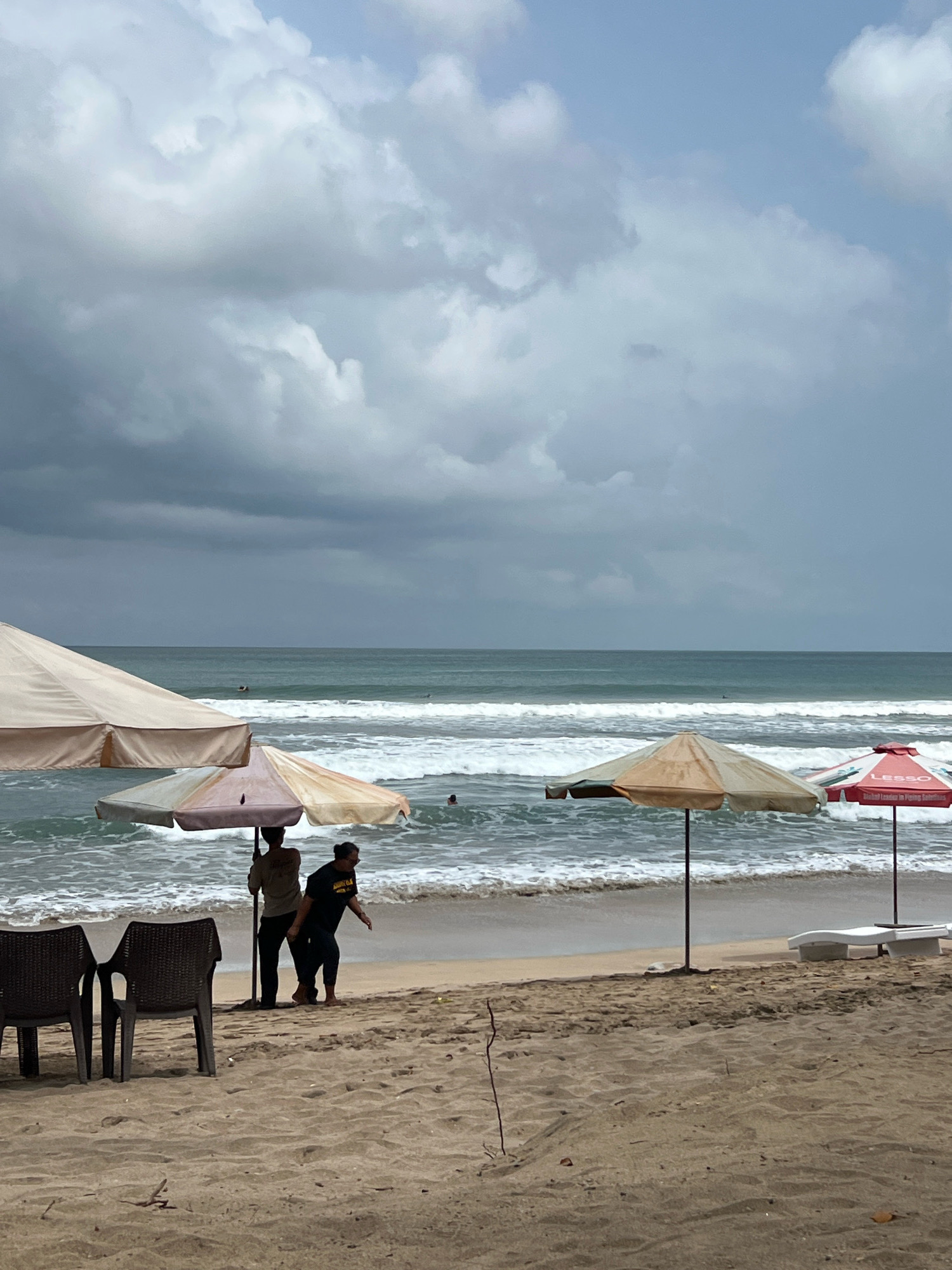 This is a vertical, medium-shot photograph taken from a beach in Kuta, Indonesia. The sky is overcast with heavy, dark clouds, suggesting an impending storm. The ocean is a turbulent teal color with white waves crashing on the shore. Two people are standing under a large, beige beach umbrella. One person, wearing a light-colored shirt, is looking away from the camera. The other person, wearing a black t-shirt with a graphic print and dark pants, is looking down and to the right. In the foreground, two dark brown chairs and a small table are set up on the sand. Further down the beach, two more beach umbrellas are visible: a smaller, striped umbrella and a red umbrella with white lettering. A white lounge chair is situated behind the red umbrella. Small pieces of driftwood and debris are scattered across the sandy beach. There are a few surfers visible in the water, and one person is swimming. The overall atmosphere of the image is moody due to the dark clouds.