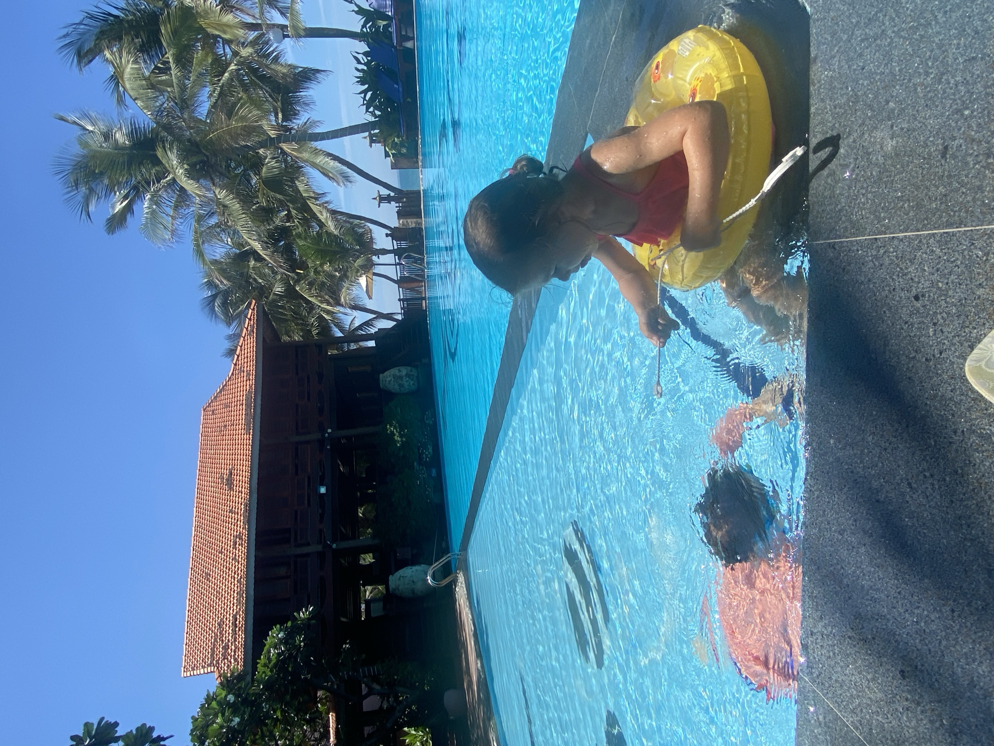 Two young children are in a bright blue swimming pool on a sunny day. In the foreground, a child wearing a pink swimsuit and a yellow inflatable ring around their waist is seated at the edge of the pool, holding a white string or rope. To the left, another child is partially submerged in the water, with their head and shoulders visible. The pool area appears to be at a resort or hotel, with a dark-tiled edge and a large, multi-story wooden building with a terracotta tile roof in the background. Palm trees are visible behind the building, and the ocean or a large body of water can be seen in the distance under a clear blue sky. The overall scene depicts a leisurely vacation activity.