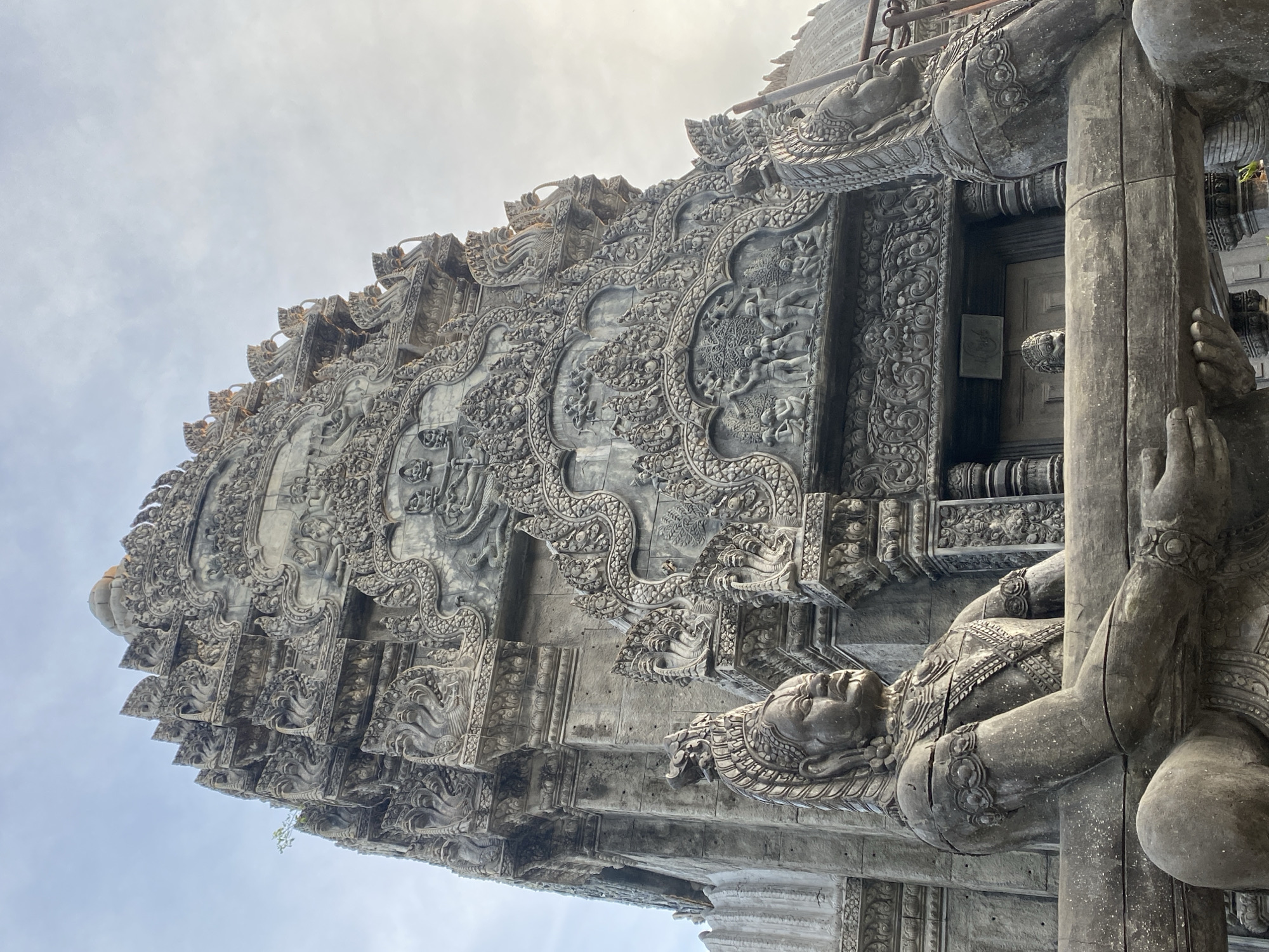 The image captures a close-up, low-angle view of an intricately carved stone structure, possibly a building or monument, located in Ban Haad Rin, Thailand. The architectural details feature extensive relief carvings depicting figures and ornate designs. The overall color palette is muted, with varying shades of gray and beige. A sculpture of a human figure is visible in the bottom right corner, seemingly supporting the structure. The sky is overcast, providing a soft, diffused lighting across the scene.