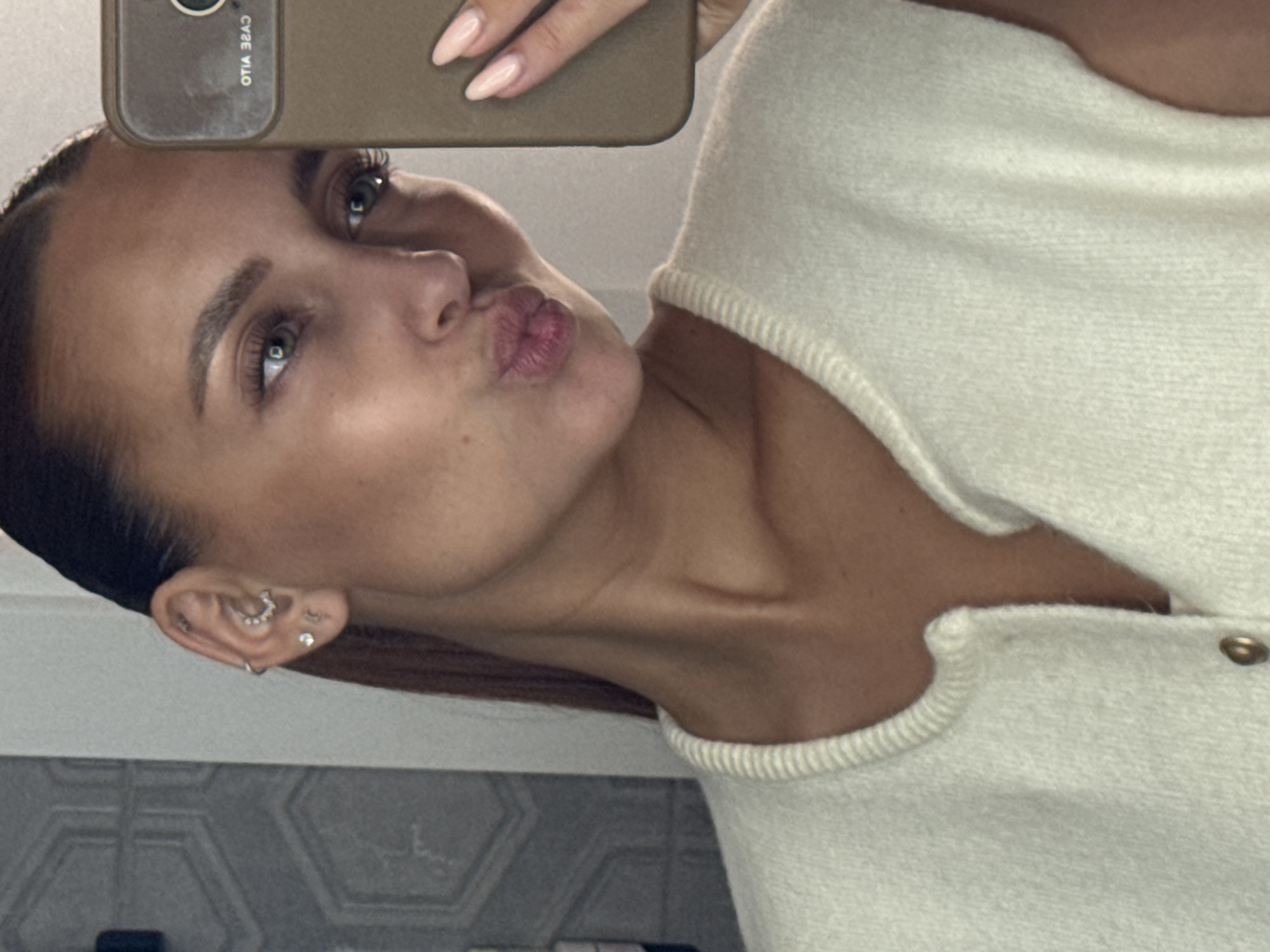 The image depicts a female individual taking a mirror selfie, captured in the city of Chaikovsky, Russia. The subject is positioned facing a mirror, holding a smartphone with her right hand, which is partially visible. Her head is tilted slightly downward, and she is making a pouting expression with her lips.
The individual has brown hair pulled back from her face, exposing her forehead and ears. Her left eye is visible, displaying a green or hazel iris with visible eyelashes and groomed eyebrows. Her right ear is visible, adorned with multiple piercings: a small silver hoop in the helix, a curved barbell or ring with small white stones in the daith, and two small stud earrings in the lobe. Her skin tone is light, exhibiting some subtle freckles on her cheek and nose area.
She is wearing a light-colored, possibly cream or off-white, sleeveless top or sweater vest. The garment features a ribbed collar and a visible gold-toned button near the neckline. The fabric appears to be knitted or textured. Her collarbone and a portion of her neck are visible.
The smartphone she is holding has a plain, possibly brown or khaki-colored, case. Text on the back of the phone case is visible and reversed due to the mirror reflection, appearing as "OTIA ЗЯАС." The nails on her right hand are manicured, elongated, and pointed, covered with a light pink or nude polish.
The background visible in the mirror reflection consists of a wall with a repeating geometric pattern in a grayish tone. The lighting appears to be internal, casting soft shadows.