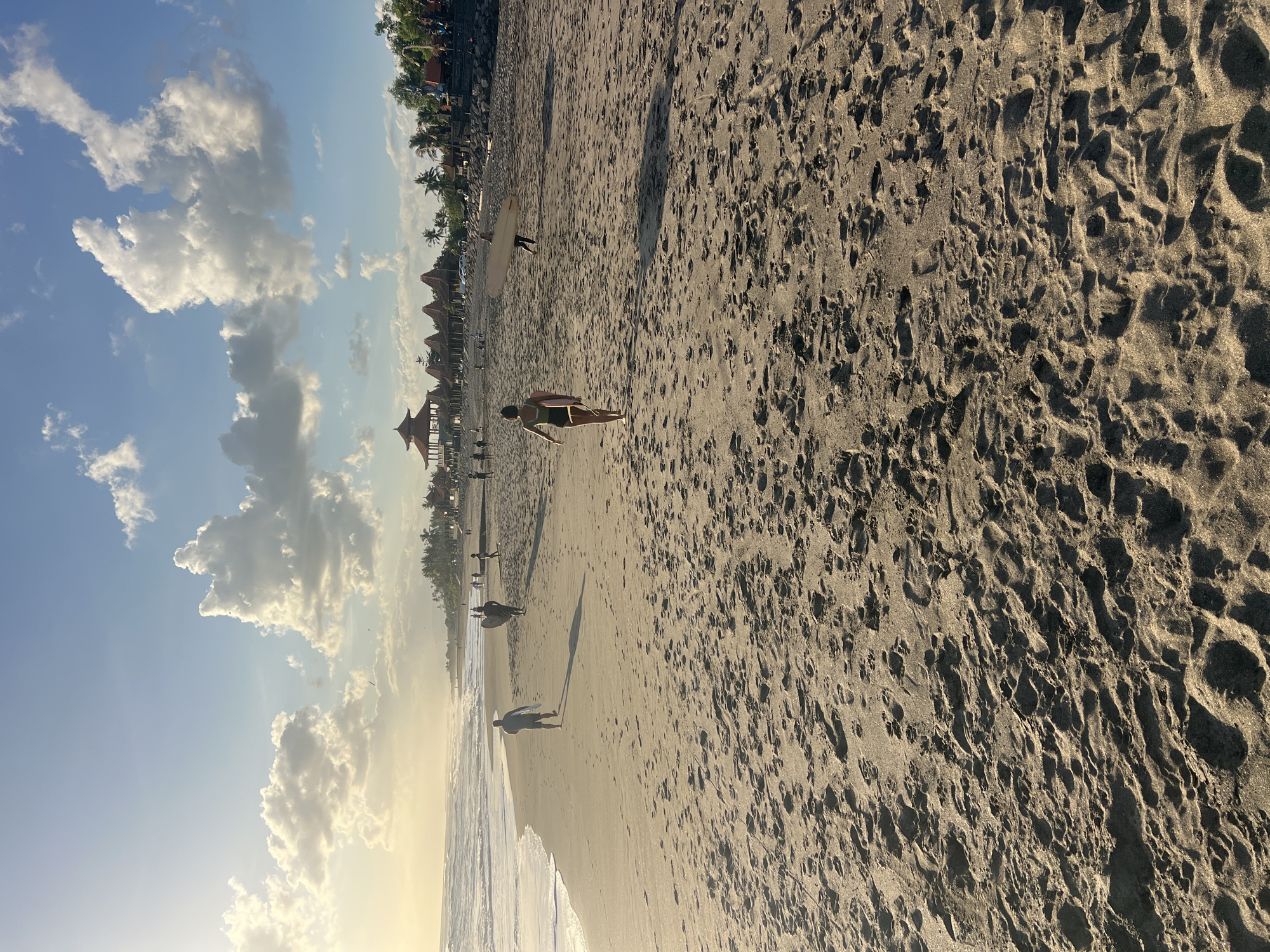 The image captures a view of a sandy beach in Kediri, Indonesia, during what appears to be late afternoon or early evening, given the warm lighting and long shadows. The beach is wide and covered in dark sand with numerous footprints. In the foreground, the sand is textured with ripples and indentations.
Further down the beach, several people are visible. A person in a dark swimsuit is walking away from the camera towards the ocean, carrying a surfboard. To the left, two individuals are walking along the shoreline. One person, seen from the back, is walking towards the ocean with a surfboard. Another person is further to the left, also walking on the sand. In the middle ground, more people can be seen scattered across the beach.
Along the distant shoreline, there are several traditional-looking buildings with distinctive peaked roofs, possibly shops or restaurants, and palm trees. The ocean is visible on the left side of the image, with moderate waves breaking on the shore. The sky is a bright blue with scattered white clouds, some of which are illuminated by the setting sun. The overall scene depicts a relaxed atmosphere at a popular beach location.