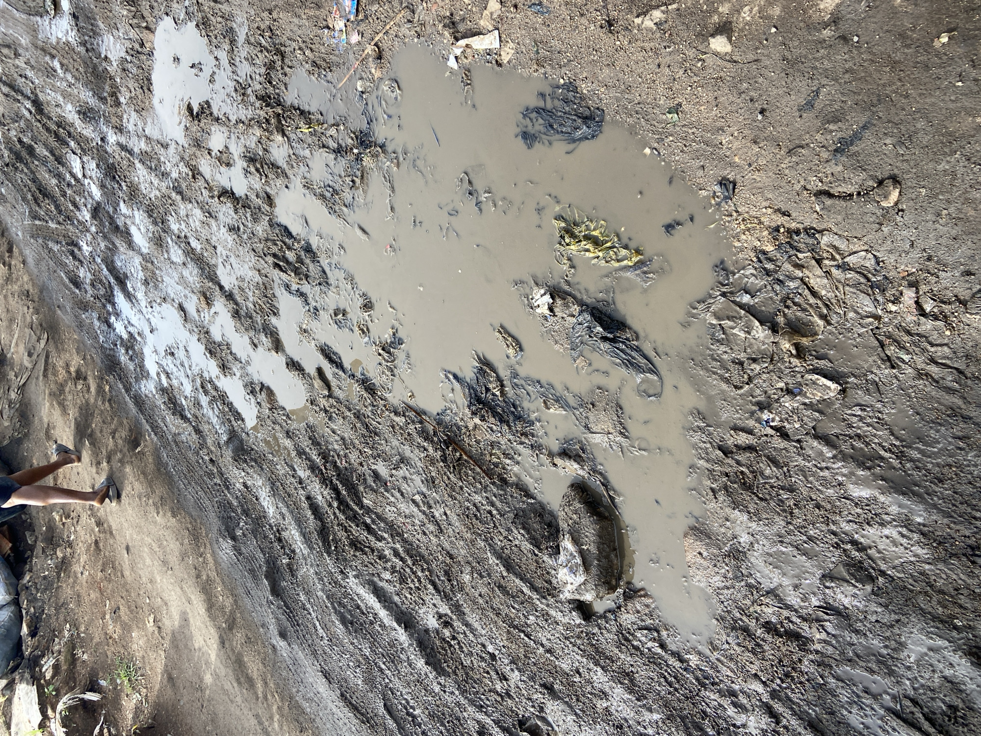 The image shows a muddy, uneven ground with a large puddle of murky water. The ground is a mix of dirt, mud, and debris, including what appears to be plastic waste. The texture of the ground is varied, with some areas appearing smooth and wet, while others are rough and packed.
In the lower left of the image, a person's legs and feet are visible, wearing dark shorts and light-colored shoes. The person appears to be standing or walking on the muddy terrain. The orientation of the image suggests it was taken from a high angle, looking down at the scene.
The lighting suggests it is daytime, with shadows visible on the ground, indicating sunlight. The overall scene depicts a neglected or damaged area, possibly an unpaved road or a construction site after rain. The presence of trash indicates a lack of proper waste management.
There is no visible text in the image. The location context is Bukuru, Nigeria.