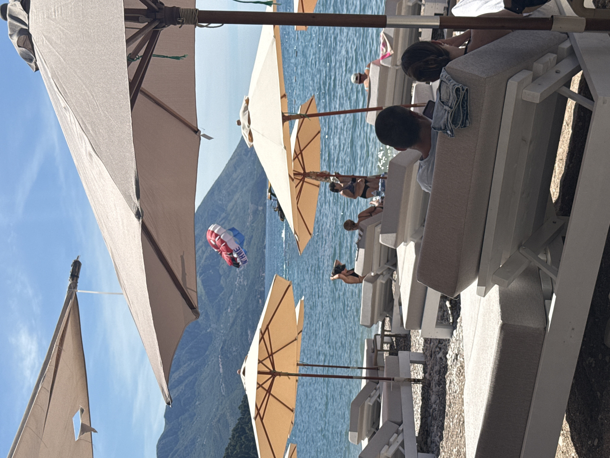 This image depicts a beach scene with several beach chairs and umbrellas set up. In the background, the Adriatic Sea is visible, with mountains in the distance. A person is parasailing in the sky, with the parachute colored red, white, and blue. The beach appears to be located in Budva, Montenegro, a popular tourist destination known for its beautiful coastline and historical old town. The scene is calm and relaxed, with people enjoying the sun and sea.