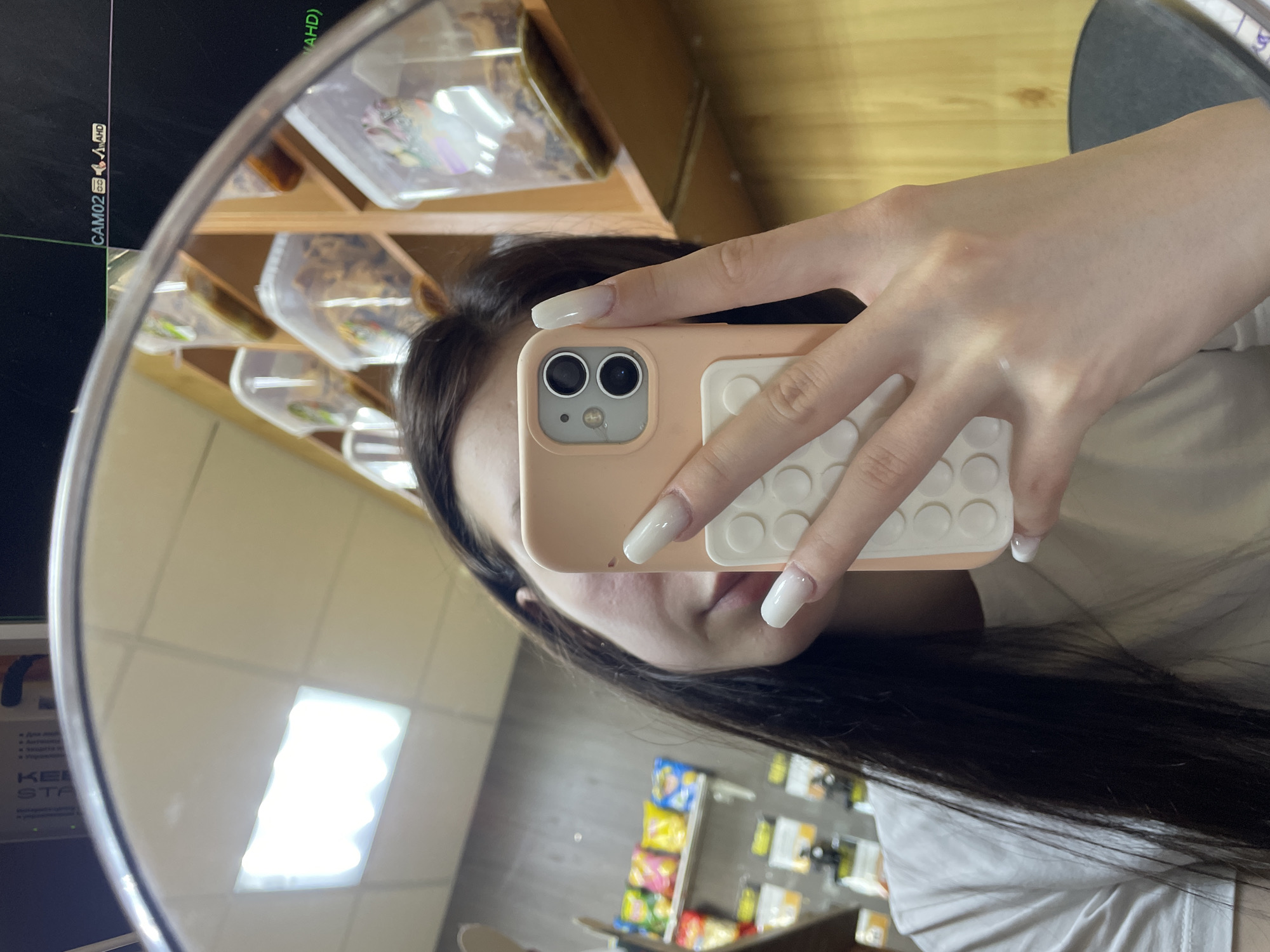 A person is taking a mirror selfie, holding a smartphone with a light peach-colored case that features a white "pop it" style circular grip on the back. Their hand, adorned with long, clear-white nails, holds the phone which obscures most of their face, revealing only parts of their dark hair and chin. They are wearing a white t-shirt.
The reflection in the circular mirror reveals an indoor setting, likely a retail space or storage area. To the upper left, a dark screen is reflected, displaying text that reads "CAM02 (AHD)", indicating a security camera feed. Below this, and continuing to the right, shelves are filled with numerous transparent plastic containers, each holding various small items. On a lower shelf to the right, colorful bags of snacks are visible. A bright rectangular ceiling light fixture is also reflected in the mirror. Partial text "KEM STA" and some Cyrillic script beginning with "Для мойки..." (For washing...) can be seen reflected, further suggesting a commercial environment. The scene is brightly lit by artificial light.