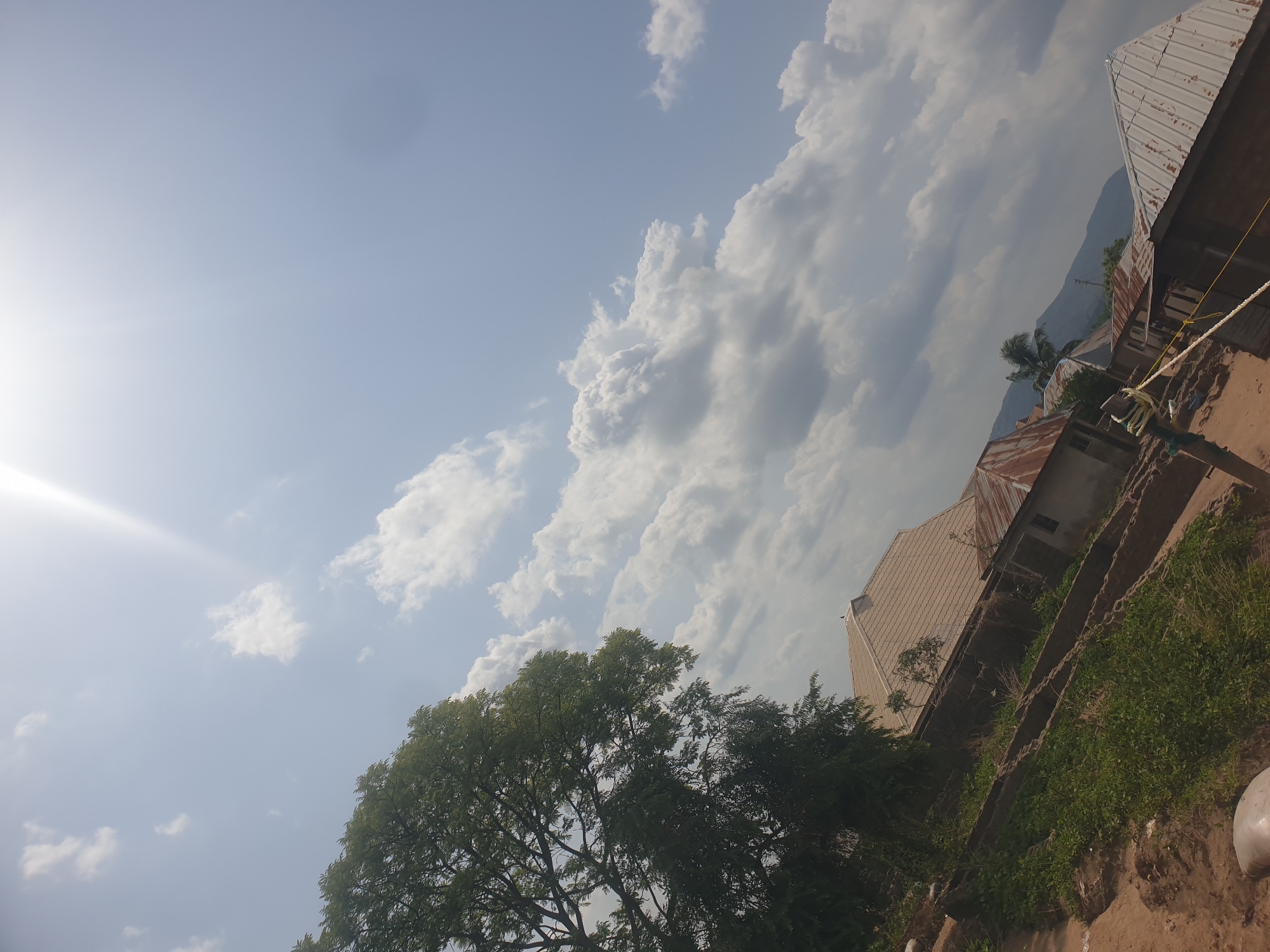 The image presents a tilted view of Zango-ndaji, Nigeria, under a sky dominated by scattered white clouds and intense sunlight. In the foreground, a tree with dark green foliage is partially visible. To the right of the frame, weathered buildings with corrugated metal roofs are situated along a dirt ground. Some vegetation grows near the buildings. The background reveals distant mountains obscured by haze. A white rope stretches across the scene.
