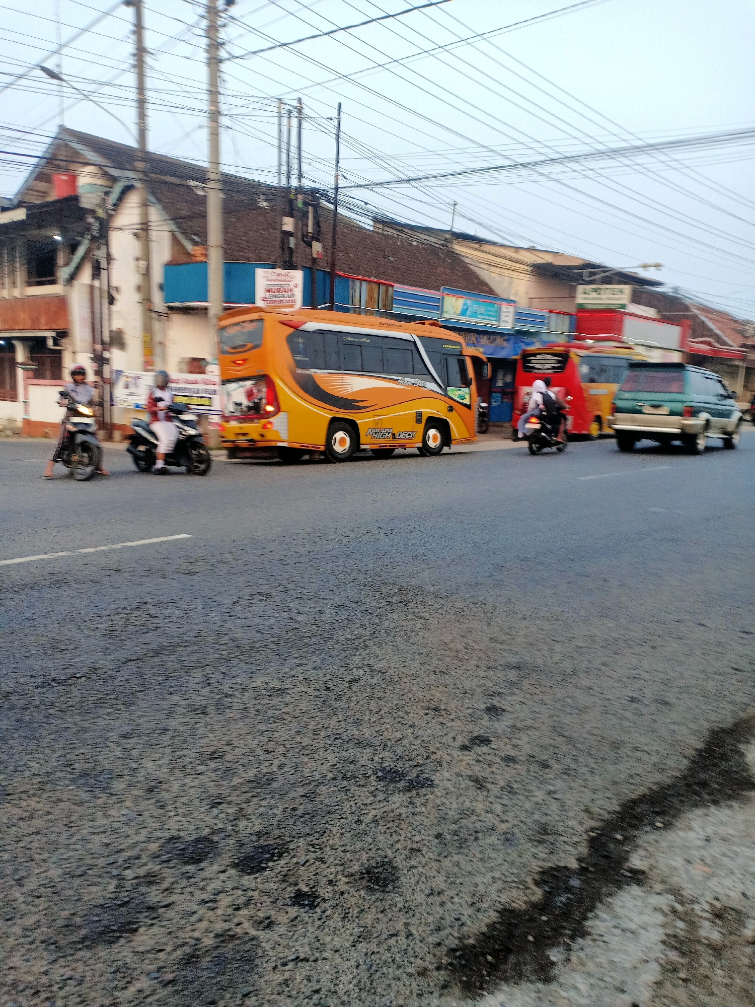 The image captures a street scene in Mojoreno, Indonesia, likely during the daytime, with overcast skies. In the foreground, a paved road with a white dividing line is visible, showing some signs of wear and dark patches, possibly oil stains.
Several vehicles are present. A bright orange and yellow bus with tinted windows is the most prominent vehicle, parked or moving slowly. Two motorcycles are on the road. One is a black motorcycle with a rider wearing a white shirt and dark pants. Another motorcycle, also black, is ridden by a person in a white shirt and helmet, with another passenger also in white. Further down the road, a red bus and a green SUV are also visible, indicating traffic.
Buildings line the side of the road. These appear to be commercial structures with signs, including one that reads "APOTEK" (pharmacy) and another with Indonesian text that is partially obscured but appears to be an advertisement. Overhead, a complex network of power lines stretches across the frame, supported by utility poles.
The overall impression is a busy, everyday street environment in a commercial area of Mojoreno.