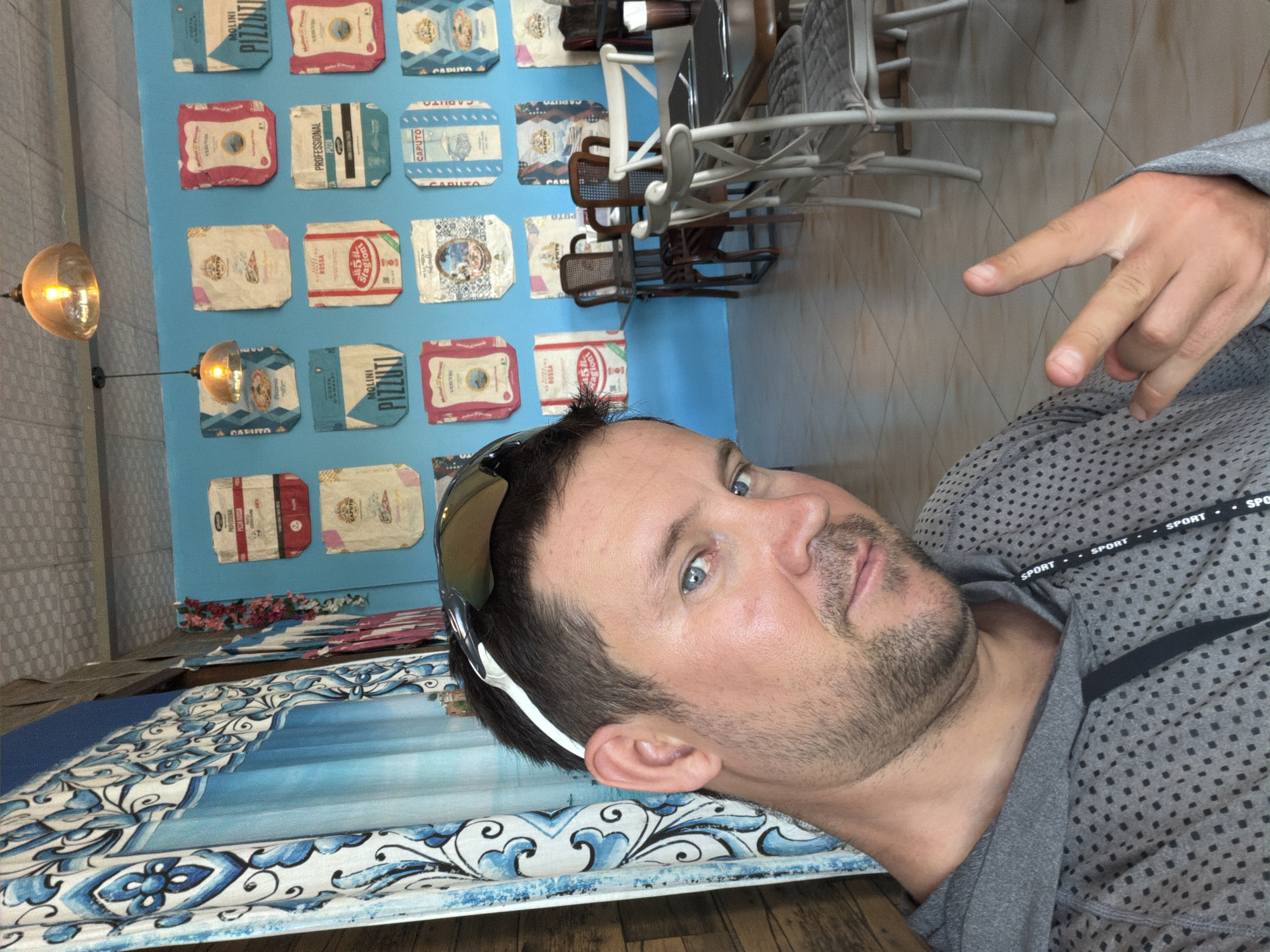 In Baan Bo Phut, Thailand, a Caucasian male with short brown hair, blue eyes, and stubble is taking a selfie indoors. He is wearing a gray hooded sweatshirt with a repeating black polka dot pattern and sunglasses pushed up on his head. He is making a hand gesture with his right hand, forming a peace sign. The background features a bright blue wall adorned with various bags, some with red and white designs and text in Italian, resembling flour sacks. A patterned blue and white curtain hangs to the left of the man. In the background, tables and chairs suggest a restaurant or cafe setting. Two pendant lights with yellowish shades hang from the ceiling. The flooring appears to be tiled.