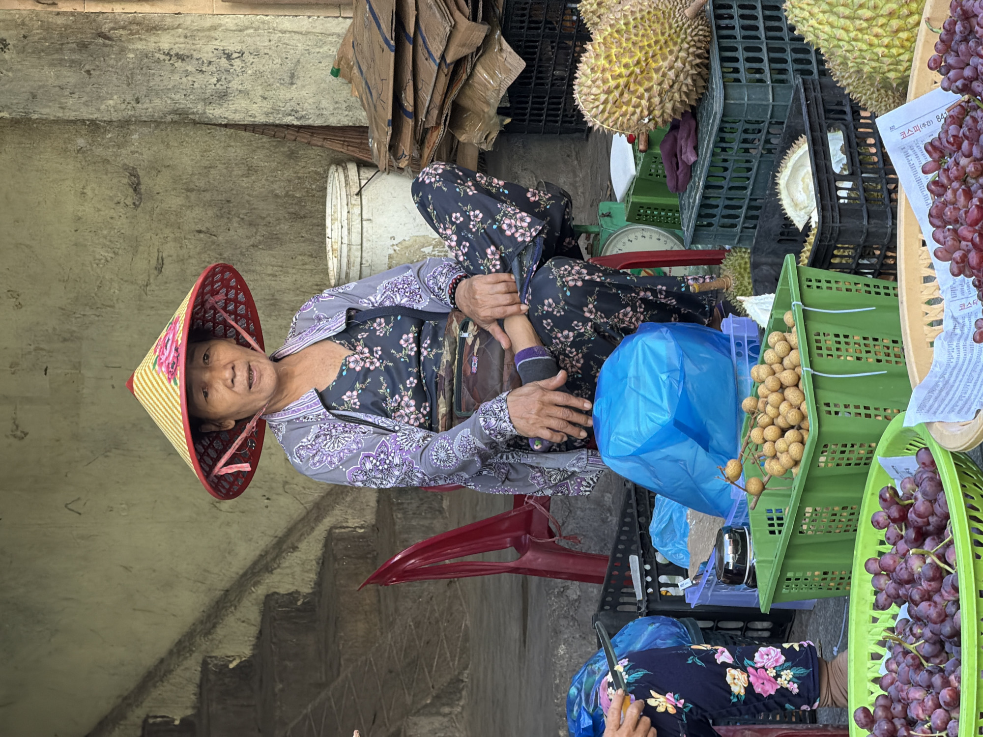 An outdoor market scene in Nha Trang, Vietnam features a woman sitting in a chair wearing a traditional conical hat, a floral shirt, and floral pants. She is positioned to the left of center, facing forward. In front of her, several baskets are filled with fruit. To her left, there are two blue plastic bags and a red plastic chair. Behind her, a white bucket and stacked cardboard boxes are visible. To the right of the woman, durian fruits and green plastic crates are present. The ground appears to be paved, and a concrete wall and stairs are in the background. The overall lighting suggests it is daytime.