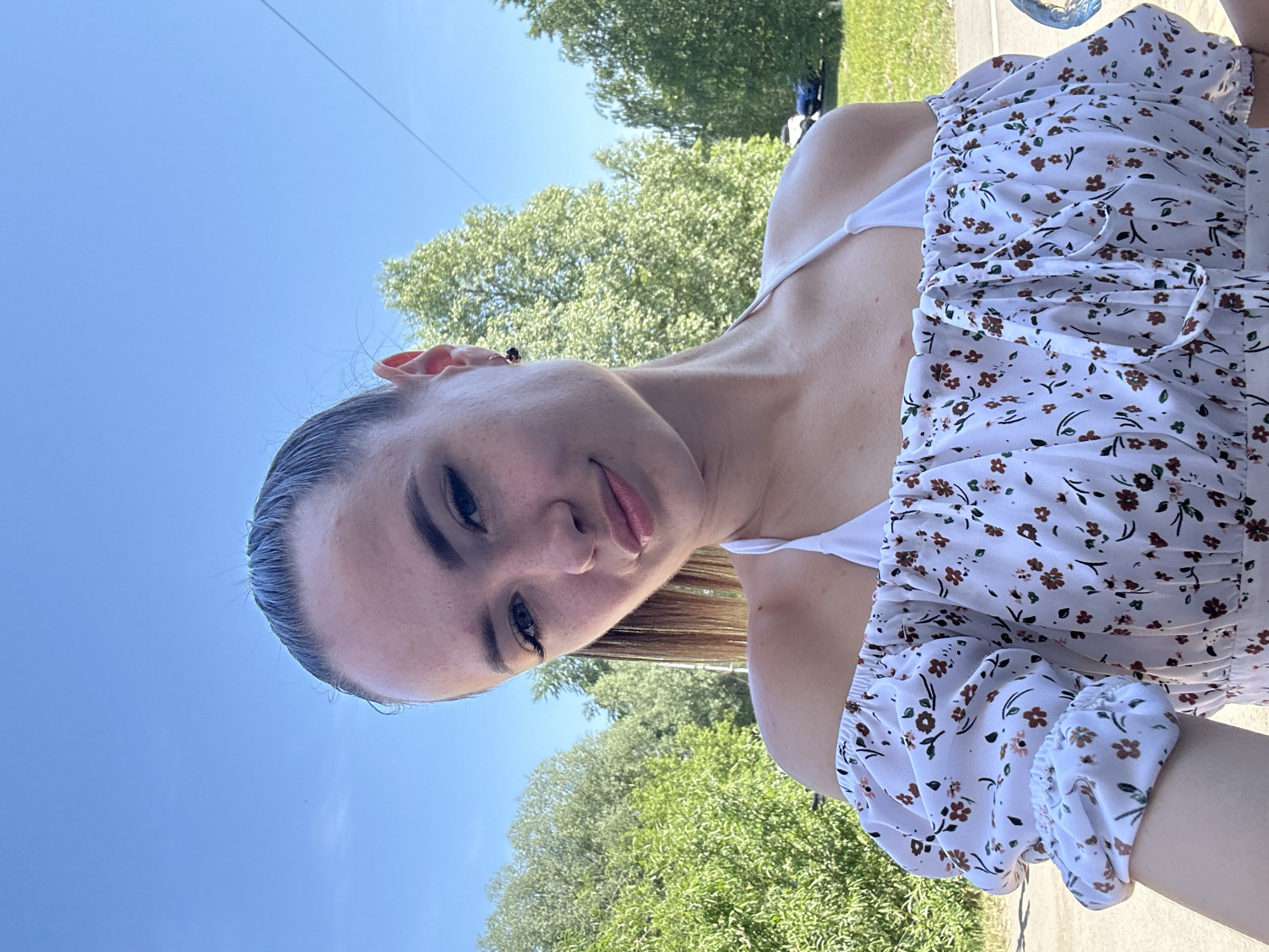 The image depicts a head-and-shoulders selfie of a young adult female in an outdoor setting in the city of Unknown, Unknown. The subject has dark hair pulled back into a high ponytail, and she wears small dark stud earrings. She looks directly at the camera with a subtle smile. Her attire consists of a white off-the-shoulder top with puffed sleeves and an elasticized neckline, patterned with small brown and light pink floral designs and green leaves. A white halter-style top is visible underneath her off-shoulder garment. The background features a clear, bright blue sky above dense green foliage from trees and bushes. A paved surface is partially visible on the far left, and a grassy area with two partially visible vehicles (one light-colored, one dark-colored) is discernible in the far right midground. The scene is illuminated by bright daylight.