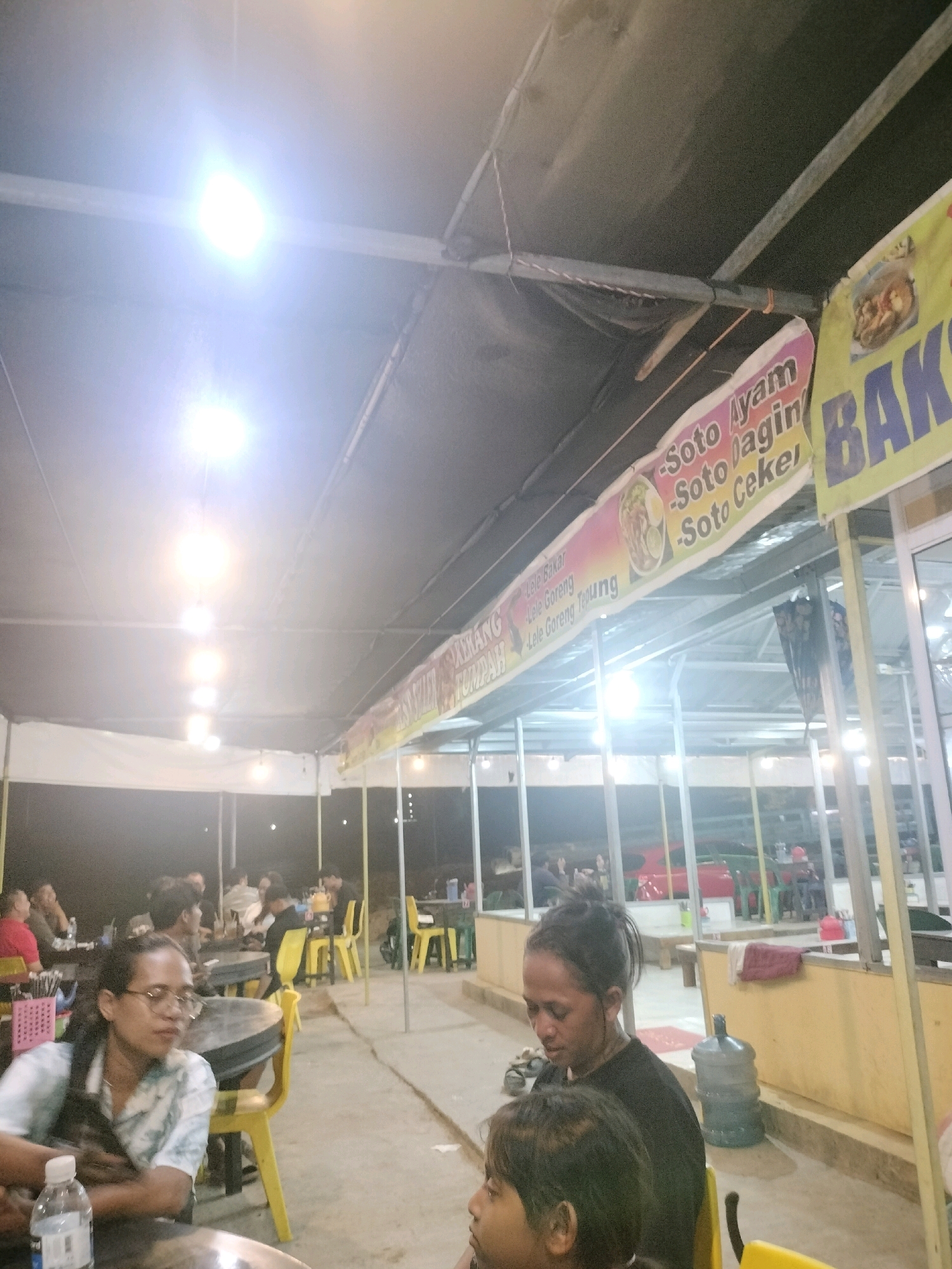 The image depicts an outdoor dining area, likely a street food stall or a casual restaurant, at night in Batam, Indonesia. Bright overhead lights illuminate the scene, casting a glare on the dark, draped ceiling. Several people are seated at tables, some engaged in conversation or eating. Yellow plastic chairs are prominent around the tables.
Banners with text in what appears to be Indonesian are hung overhead. One banner is yellow and features the word "BAKSO" in large blue letters, suggesting it is advertising meatballs. Another banner, with a pink and yellow gradient, lists food items like "Soto Ayam," "Soto Daging," and "Soto Ceker," indicating it's a menu for various types of soto (Indonesian soup). There are also signs mentioning "Lele Bakar" (grilled catfish) and "Lele Goreng" (fried catfish).
In the foreground, two women are seated at a table. One woman, wearing glasses and a patterned shirt, is looking to her left. Next to her, another woman with her hair pulled back is looking down. A water bottle is visible on the table in front of the woman with glasses. In the background, other patrons are visible, and a large water dispenser is seen to the right. The overall atmosphere appears to be relaxed and casual, typical of an evening dining establishment in Indonesia.