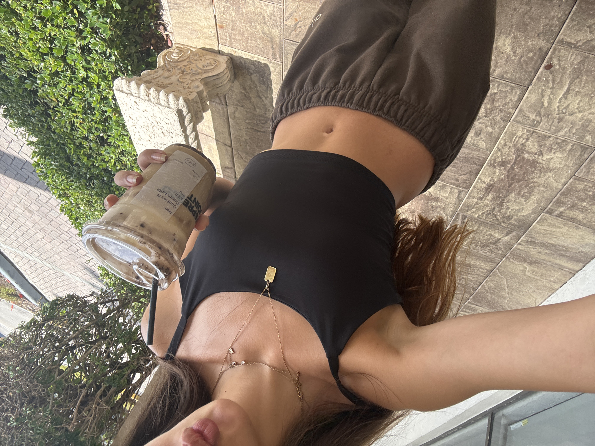 A slightly downward-angled, front-facing image captured outdoors in Boca Raton, United States of America, depicts an individual from the upper torso to the upper thighs. The individual has tan skin and long brown hair, visible on the right side. They are wearing a black spaghetti strap crop top, exposing the midriff and navel, paired with dark brown elastic-waist sweatpants featuring a white logo on the left thigh. Multiple layered gold-toned necklaces adorn the individual's neck: a choker-style necklace with small clear stones, a shorter chain with a small charm, and a longer chain with a rectangular gold tag pendant. The individual's lips are partially visible at the upper right, slightly parted. In their right hand, they hold a clear plastic cup containing an iced 'Cookies N Cream Latte' from 'Espresso Point', featuring a black straw. The cup's label displays the name 'Ivan', 'ICED', 'REG', and '2 of 4'. The ground is paved with large, textured grey-brown tiles. An ornate, light-colored stone baluster is positioned to the right. The background features dense green foliage from hedges and trees with visible branches, a paved area, and a partial view of a windowed building to the left. The scene is brightly illuminated by natural sunlight.