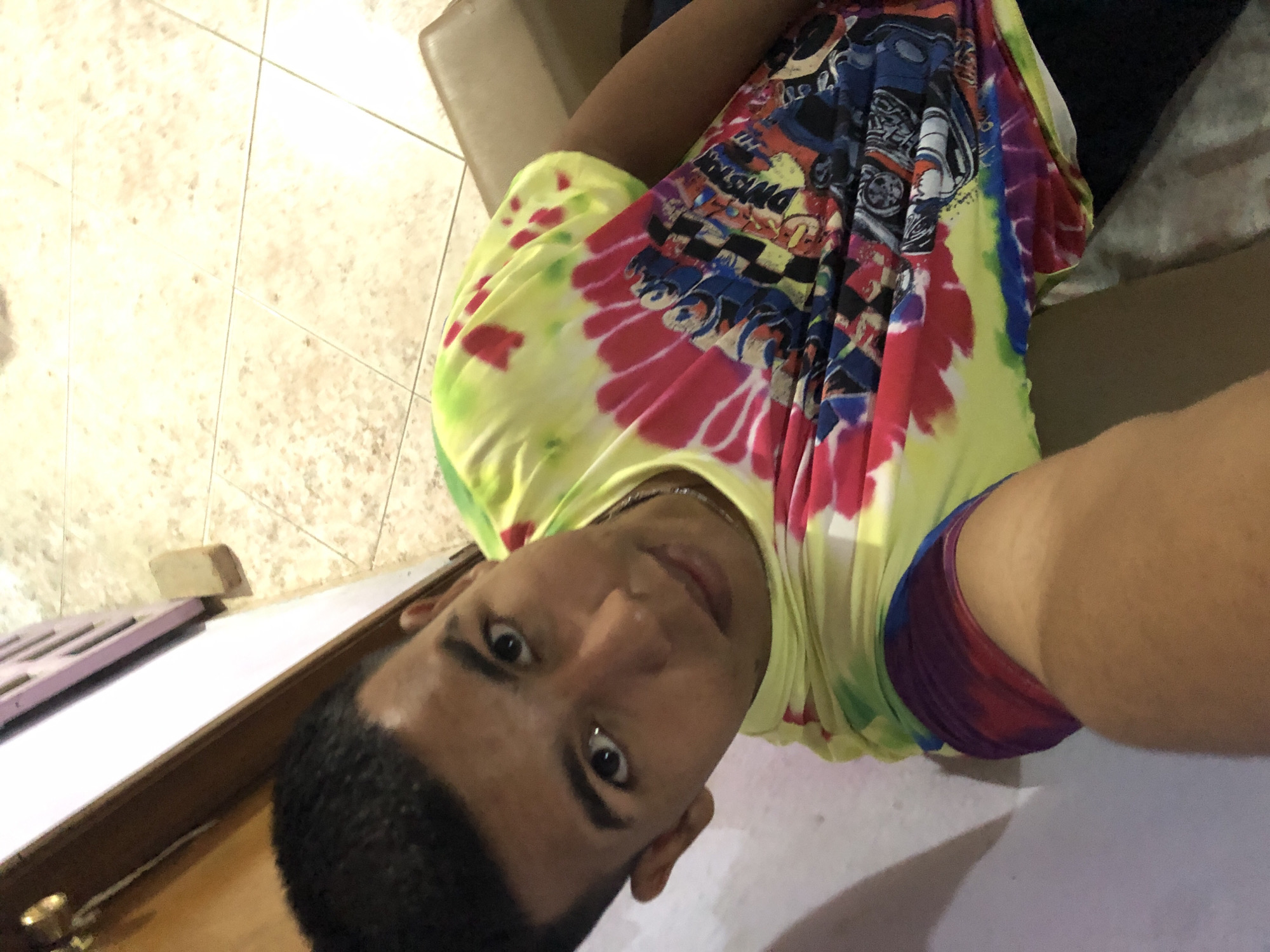 A young man with dark hair is taking a selfie. He is wearing a brightly colored tie-dye shirt with racing car designs. He has a slight smile and is looking at the camera. The background is a tiled floor and a piece of furniture. There is no information in the image to determine its location in Altagracia, Venezuela.