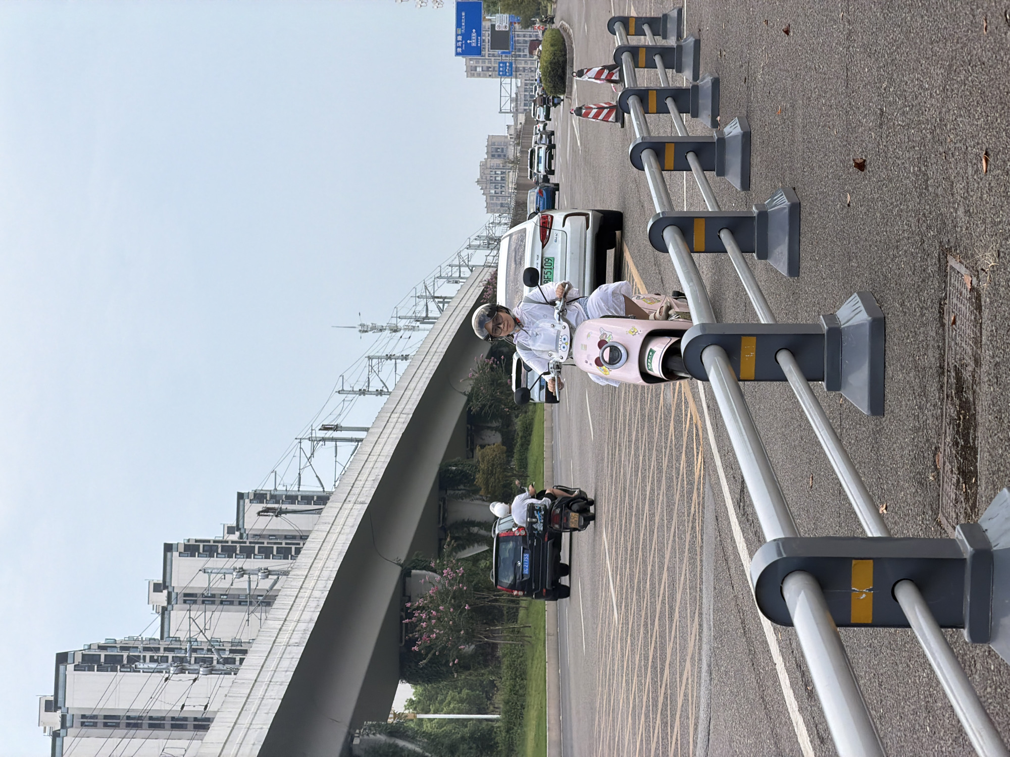 A woman rides a pink electric scooter on a cloudy day in Jiangjin, China. She is wearing sunglasses, a helmet, and a light-colored outfit. She is on a multi-lane road with other vehicles, including cars and motorcycles. A large overpass with train tracks runs above the road. The road is lined with a grey metal railing with yellow accents. In the background, there are apartment buildings and a road sign. The scene appears to be a typical urban environment in China.