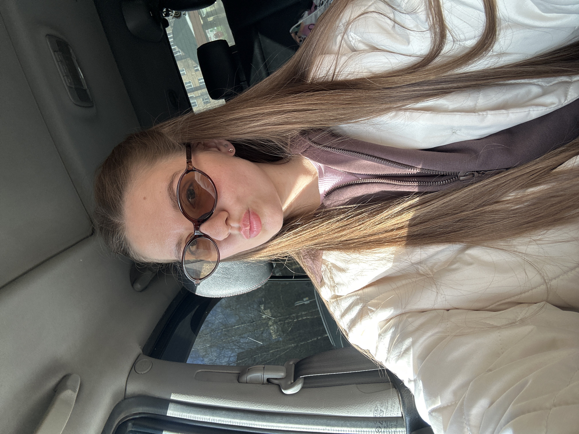 A young woman with long brown hair is wearing glasses and a white jacket. She is inside a vehicle. The window shows a building in Klin, Russia.