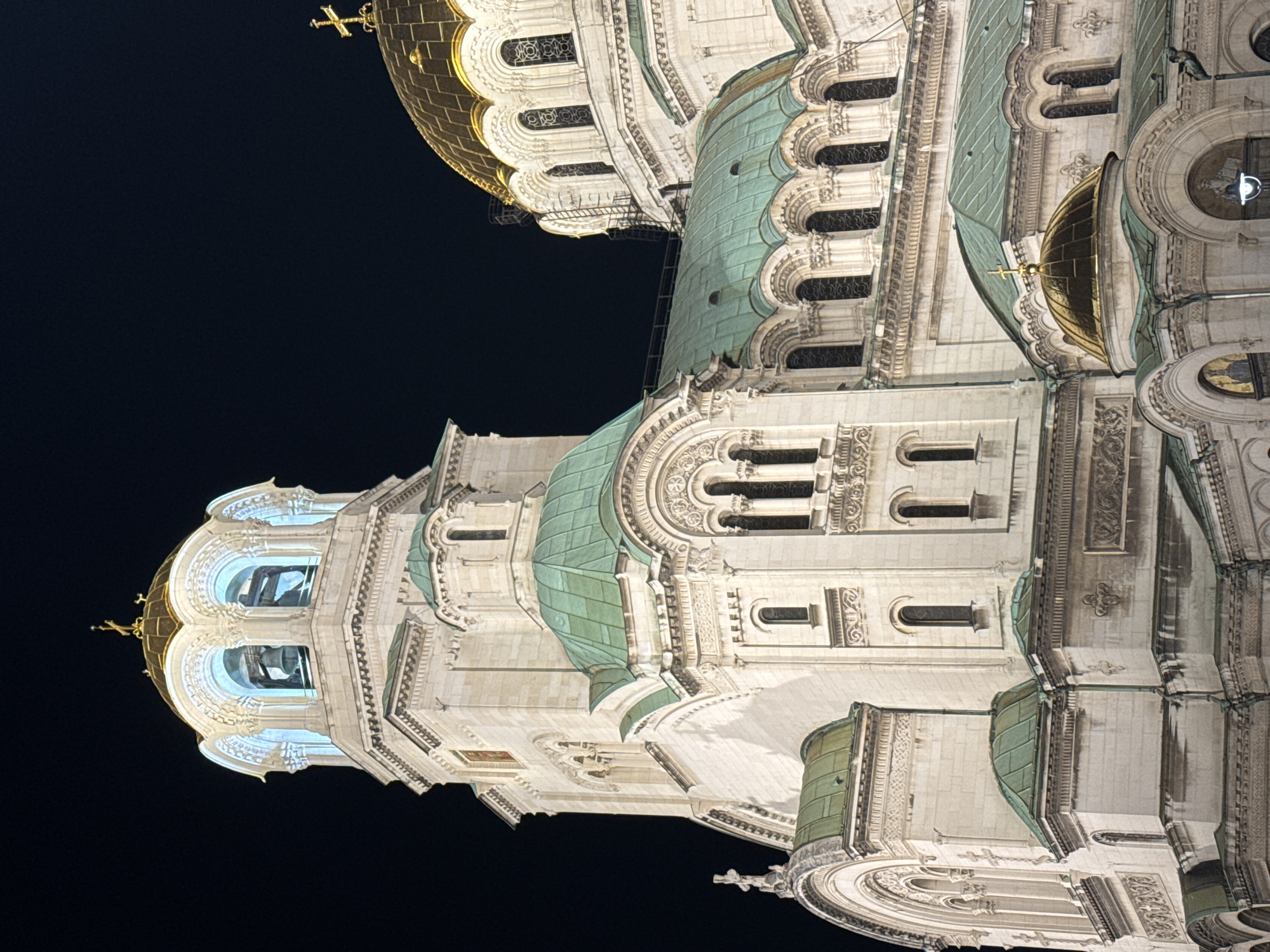 The image shows the Alexander Nevsky Cathedral in Sofia, Bulgaria, at night. The cathedral is illuminated, highlighting its architectural details, including domes, arches, and windows. The sky behind the cathedral is dark. The cathedral's exterior is primarily white stone. Some of the domes are covered in gold and some are covered in green tiles. The cathedral is richly decorated with carvings and ornamentation. No people are visible in the image.