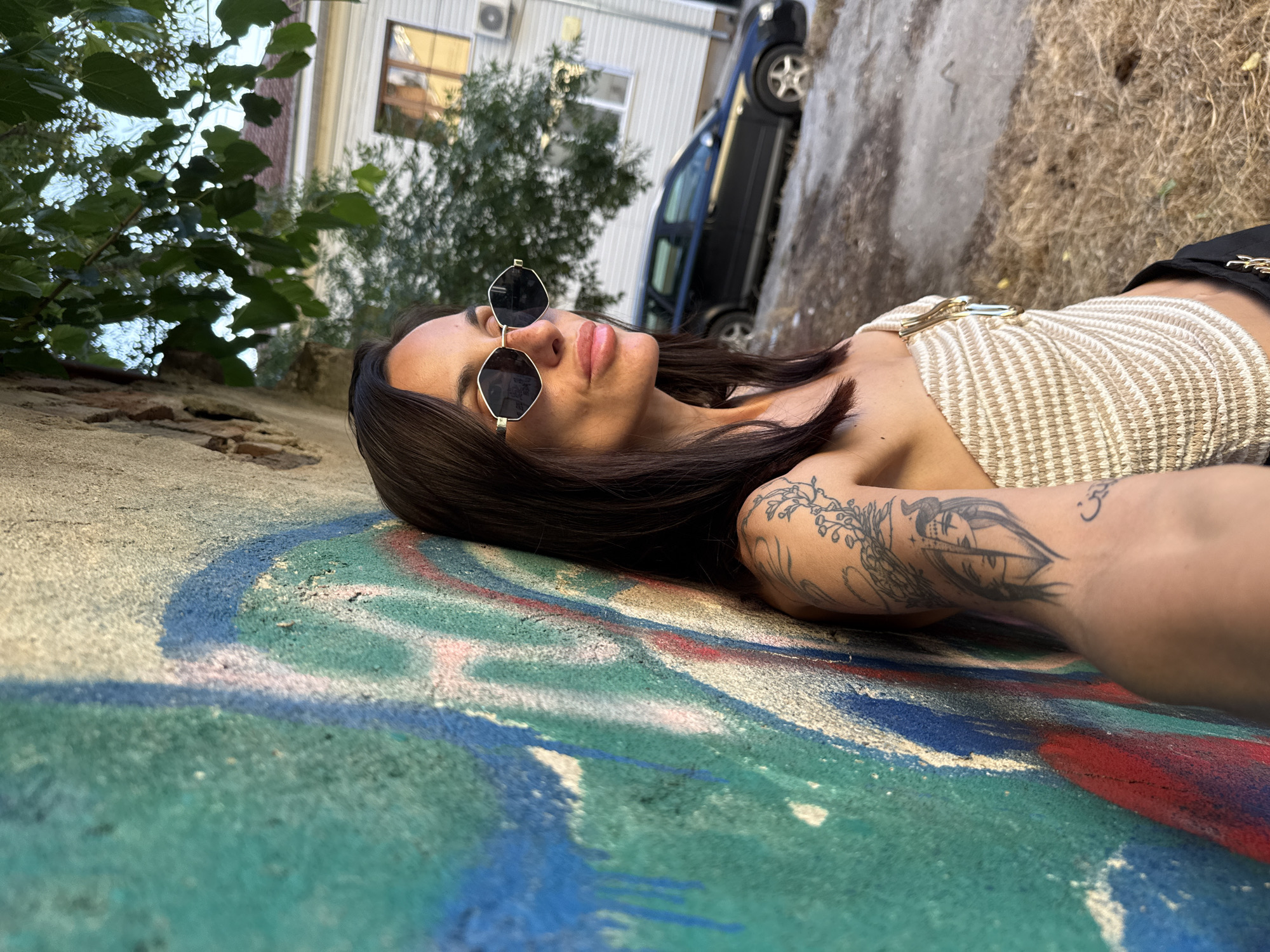 The image shows a woman leaning against a graffiti-covered wall in an urban environment. The wall, which is textured and appears to be crumbling in some areas, features vibrant spray-painted designs in blue, green, and red. The woman has long, dark hair and is wearing sunglasses, a strapless top, and black pants. She has tattoos on her arm, including a detailed design of a face. Behind her, a black car is parked on a dirt road, and a building with windows and an air conditioning unit is visible. Trees with green leaves hang over the scene. The lighting suggests it is daytime, possibly late afternoon. The location is identified as Pleven, Bulgaria.