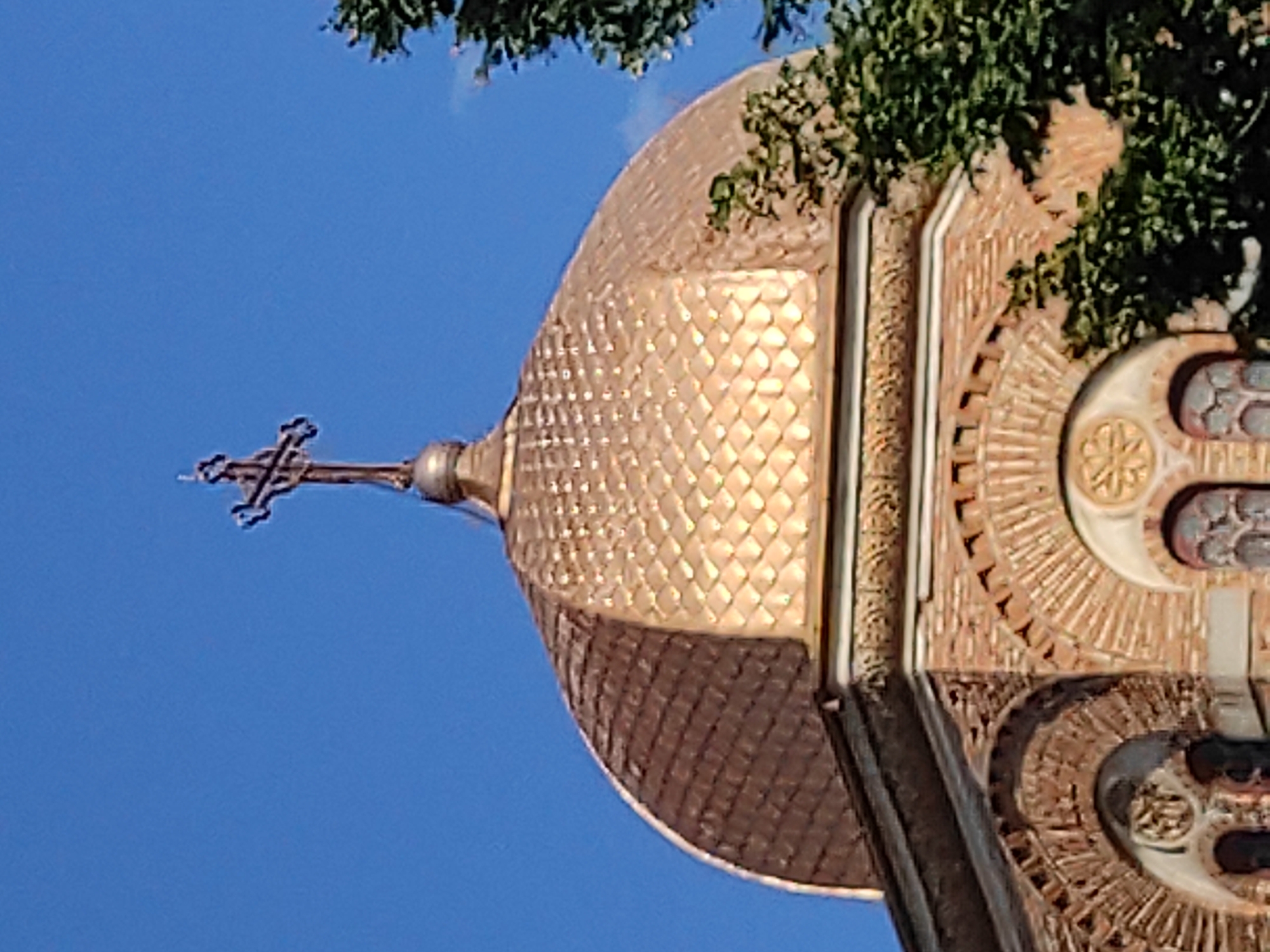 The image shows the golden dome of a church, topped with a cross. The dome is covered in diamond-shaped tiles. Below the dome, the church is constructed of brick with arched windows. There is a tree to the right of the dome. The sky is a clear blue. This is likely the dome of Saint Andrew's Church in Constanța, Romania, as it features a prominent golden dome and is a well-known landmark in the city.