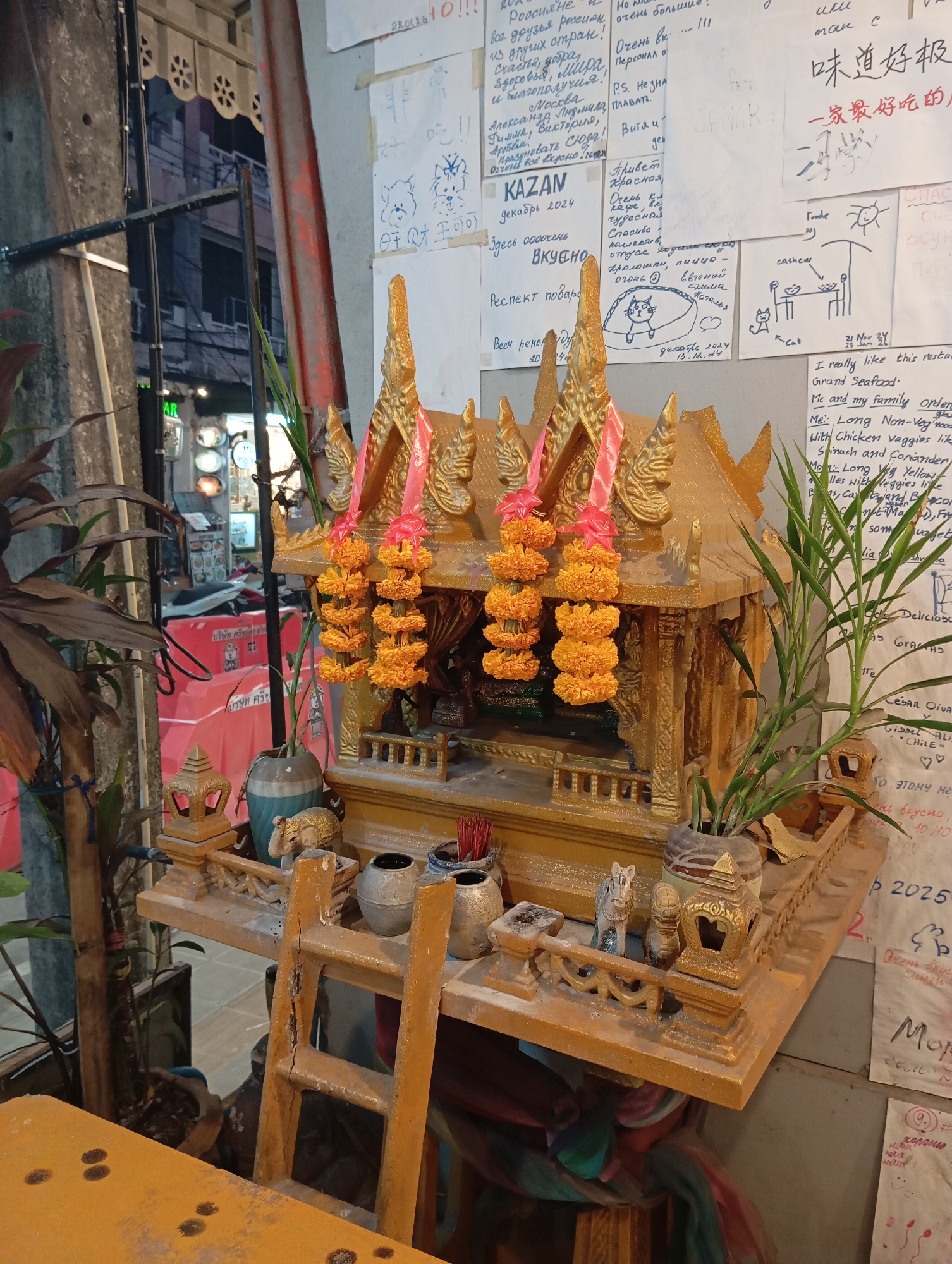 In Karon, Thailand, an outdoor shrine, possibly a spirit house, is set up on a wooden platform supported by a ladder. The shrine is ornate and golden, adorned with vibrant orange marigold garlands. On the platform, various items are arranged, including small decorative objects like elephants and lanterns, as well as incense sticks in holders and two ceramic vases. To the right of the shrine, a potted green plant adds a touch of nature. The background shows a slightly blurred view of a street with buildings, suggesting an urban or commercial setting. The overall scene is a blend of cultural elements and everyday surroundings.
