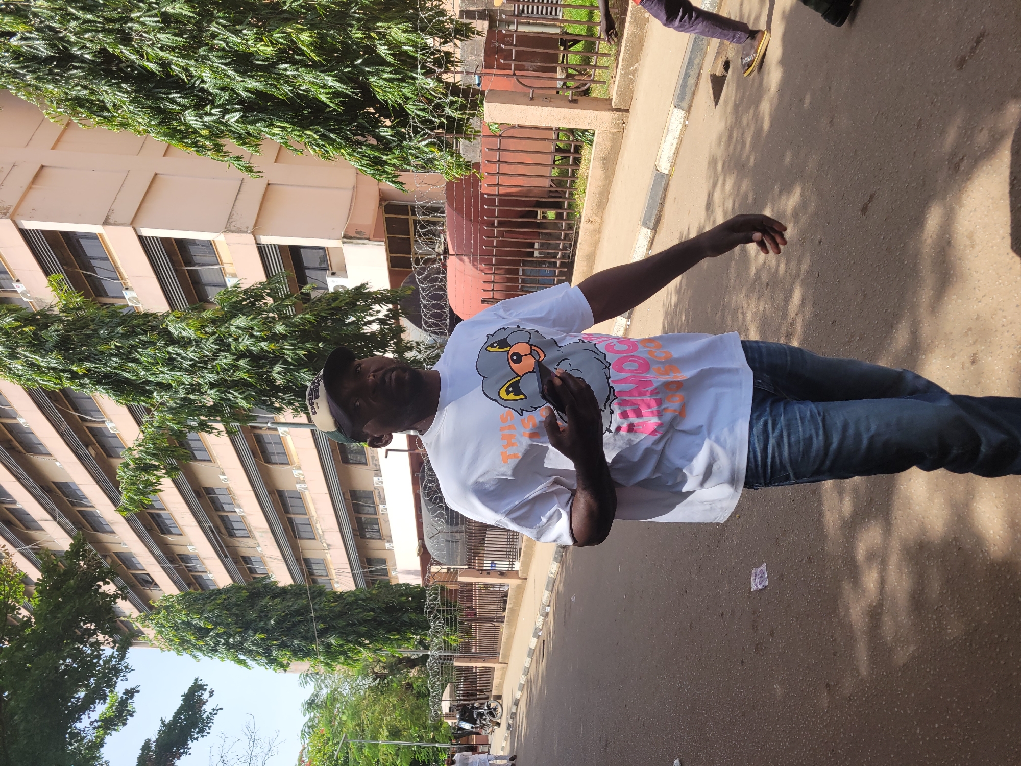 A man stands prominently on a paved street in an urban setting, likely in Abuja, Nigeria, under bright daytime sunlight. He has dark skin, is wearing a white short-sleeved t-shirt, blue jeans, and a cap that is partially visible. His left arm is extended outwards to his side, while his right hand holds a mobile phone.
The man's white t-shirt features a distinctive graphic design: a grey, cartoon-style bird or duck-like character with an orange beak, wearing what appears to be a yellow crown or hat. Below this graphic, the words "THIS IS" are visible in orange lettering, followed by less distinct text in pink.
Behind the man, a multi-story building with a light beige or peach facade and numerous windows stretches across the background. Several air conditioning units are visible beneath the windows. Dense, dark green trees or bushes line the street and partially obscure the building. A metal fence, topped with barbed wire, runs along the perimeter of the property, separating it from the street. Behind the fence, what appears to be an electricity transformer can be seen.
The street is paved, with a curb featuring grey and white sections. Strong sunlight casts sharp shadows on the ground, indicating a clear, sunny day. The scene suggests a typical urban environment, possibly a street adjacent to an office building or a residential complex. No specific activity or event beyond the man standing and holding his phone is discernible, and his expression appears neutral.