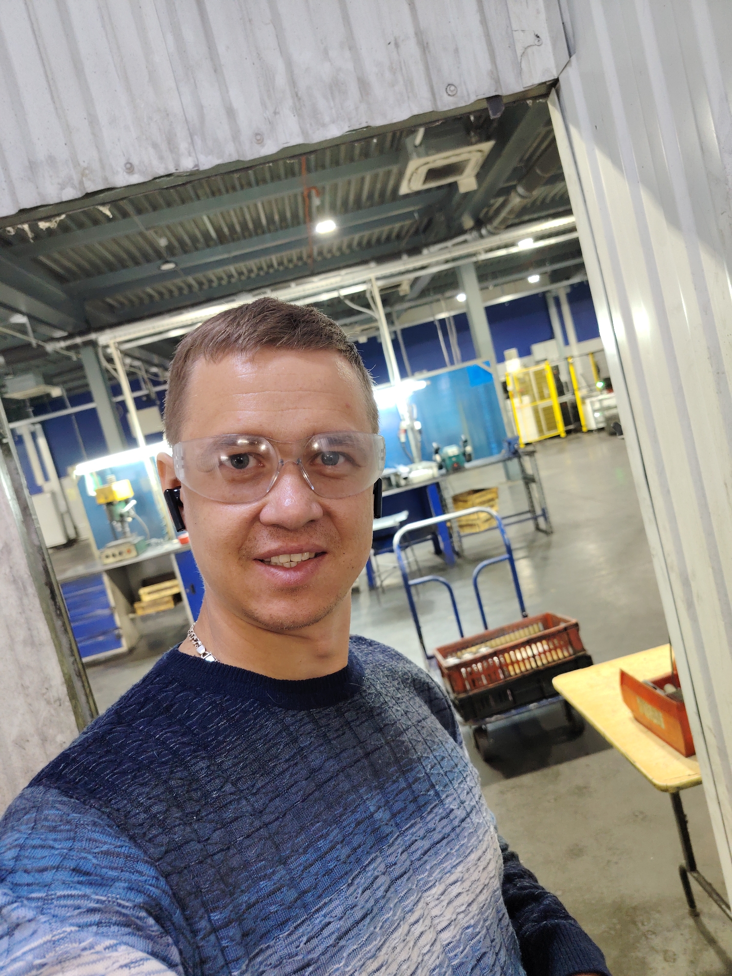 A man, appearing to be in his 30s or 40s, with short light brown hair, is captured in a selfie taken indoors. He wears clear safety glasses and dark earbuds in both ears. A faint smile is on his face, and he has a silver-colored chain necklace around his neck. He is dressed in a textured, gradient-patterned sweater that transitions from dark blue at the top to lighter blues and white towards the bottom.
The man is situated in what appears to be an industrial workshop or factory setting, likely in Noviy, Russia. The background reveals a spacious area with a high ceiling featuring exposed metal structures, pipes, and overhead fluorescent lighting. The floor is concrete. On the left side of the frame, behind the man, a drill press on a workbench is visible. Further back, there are work areas with tables, blue-painted walls, and various pieces of machinery, some enclosed with yellow safety barriers. In the mid-ground, a blue two-wheeled cart is visible, along with a red plastic crate resting on a black wheeled trolley. Another small, light-colored table with an orange container sits on the far right. The environment suggests an active workspace where protective gear, like the man's safety glasses, is appropriate. There is no visible text in the image.