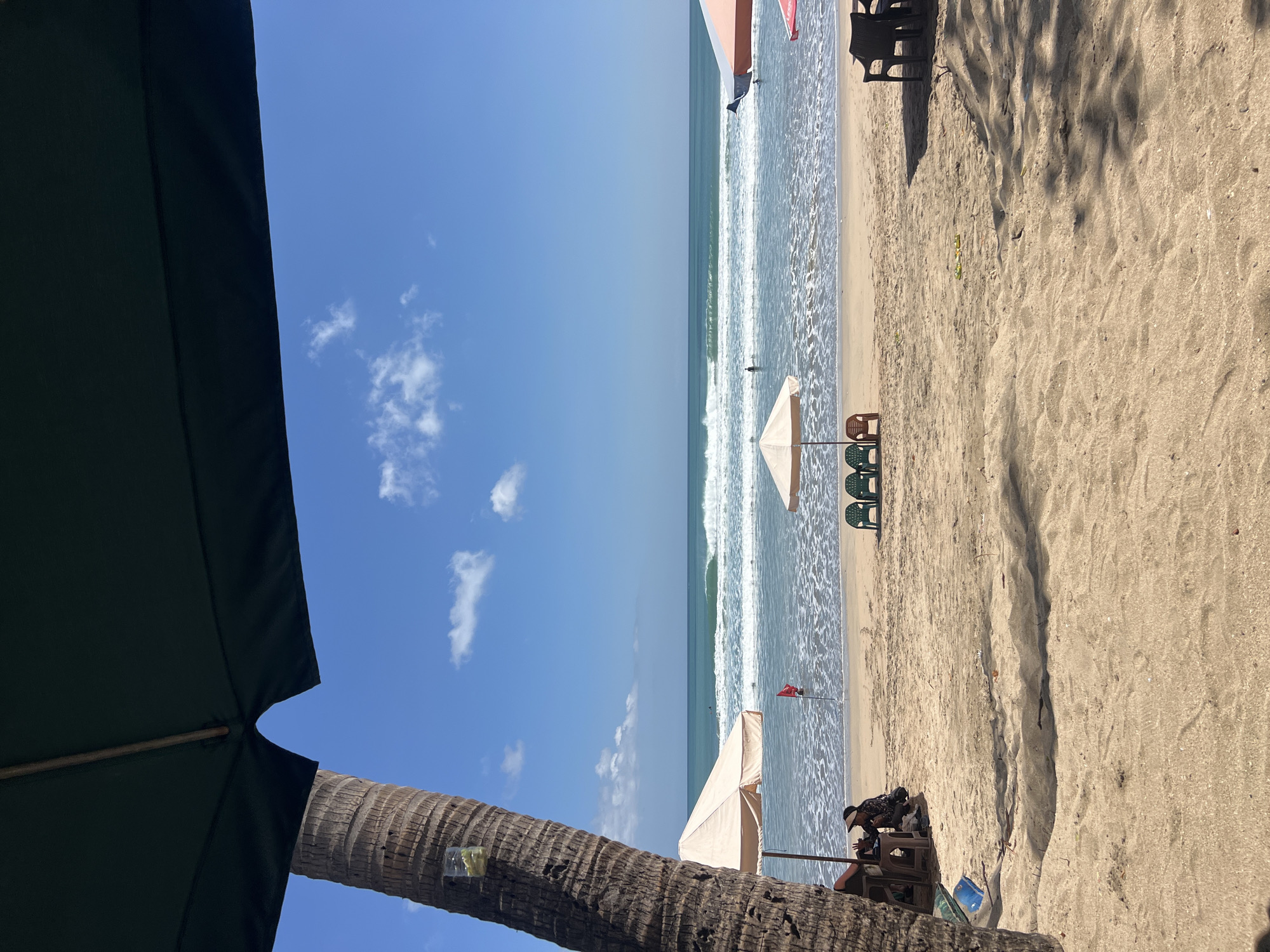 This is a daytime, eye-level, horizontal, outdoor photograph taken on the beach in Kuta, Bali, Indonesia. The scene features a sandy beach with the ocean in the background.
In the foreground, a large palm tree trunk is visible on the left, with a dark umbrella overhead, obscuring the top portion of the frame. To the left of the palm tree, a woman wearing a hat is seated at a small table under a tan umbrella. She appears to be interacting with someone out of frame or a tablet. Further to the right, two green beach chairs and one brown beach chair are positioned in front of a small, light-colored umbrella.
The ocean displays moderate waves breaking on the shore, with white foam visible. A single individual is standing in the shallow water. Further out in the water, several other figures are visible, some of whom appear to be surfers. A red flag is planted in the sand near the water's edge. On the right side of the frame, a table and chairs are partially visible under another umbrella. The sky is blue with scattered white clouds. The sand on the beach is light brown and textured, with visible footprints and some scattered debris.