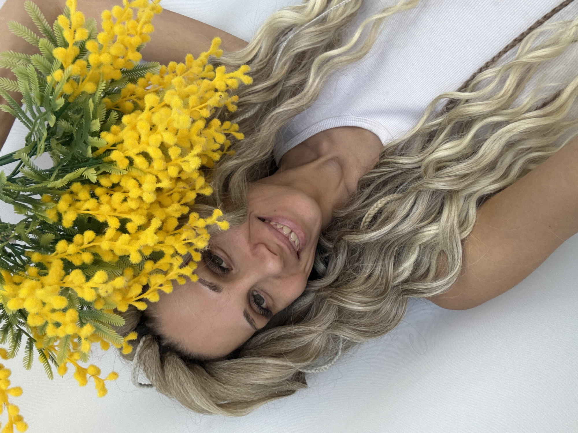 A woman lies on a white surface next to a bouquet of yellow flowers. She wears a white shirt and has long blonde hair with braids. She smiles at the camera. The location is not specified in the image.