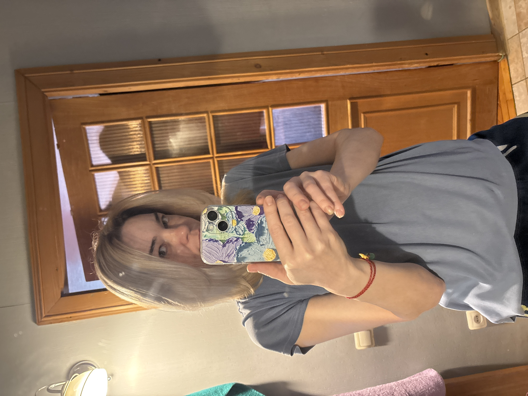 A woman is taking a selfie in a bathroom. The location is Unknown, Unknown. She is holding a smartphone with a floral-patterned case. She is wearing a gray T-shirt. There is a wooden door behind her. Below her, on the sink are a turquoise towel and a pink towel. There is a light fixture on the wall to the left.