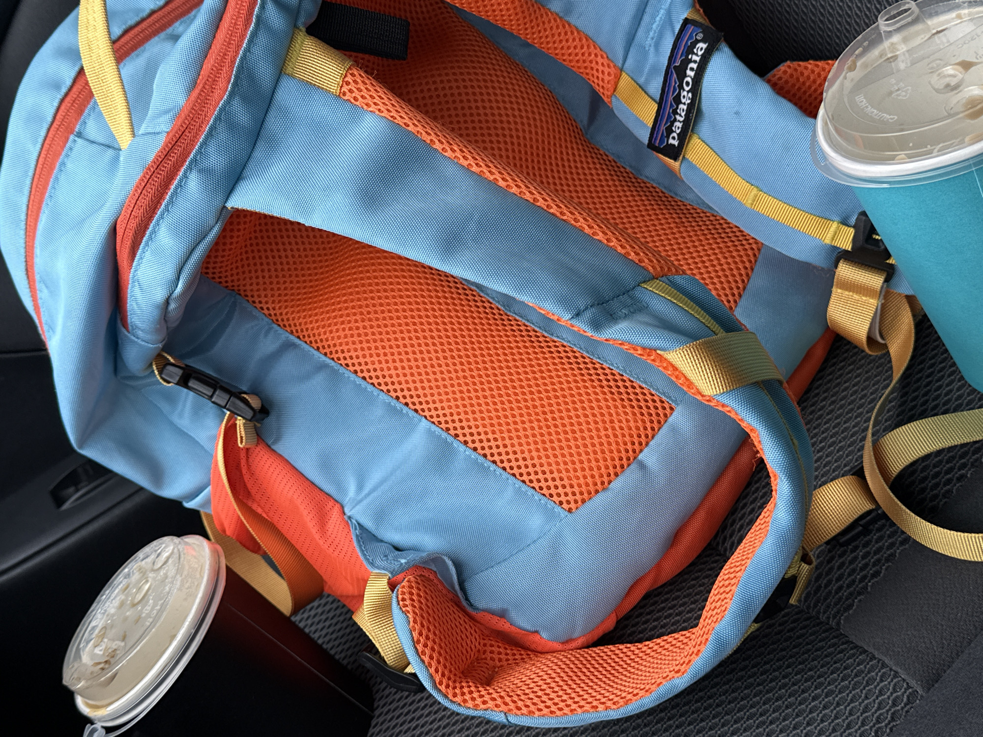 The image shows a blue, orange, and yellow Patagonia backpack resting on a dark gray, patterned surface, likely a car seat. There are two cups of iced coffee, one to the left of the backpack and one to the right. The location is not identifiable.
