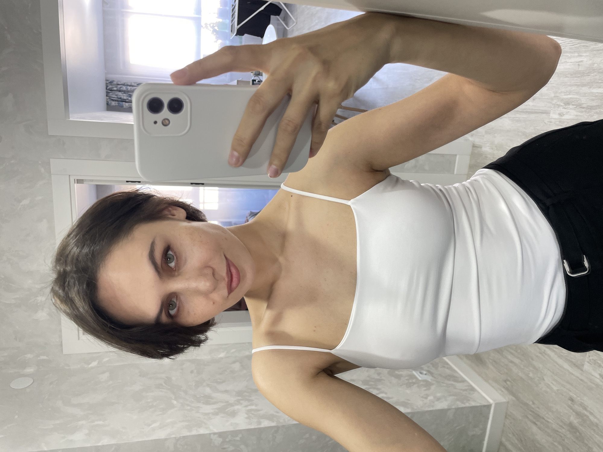 A woman is taking a selfie in a bathroom. She is wearing a white tank top and black pants. The bathroom has light grey marble flooring. The woman is holding a white iPhone. A window is visible in the background. There is no evidence to suggest this image was taken in Izhevsk, Russia.