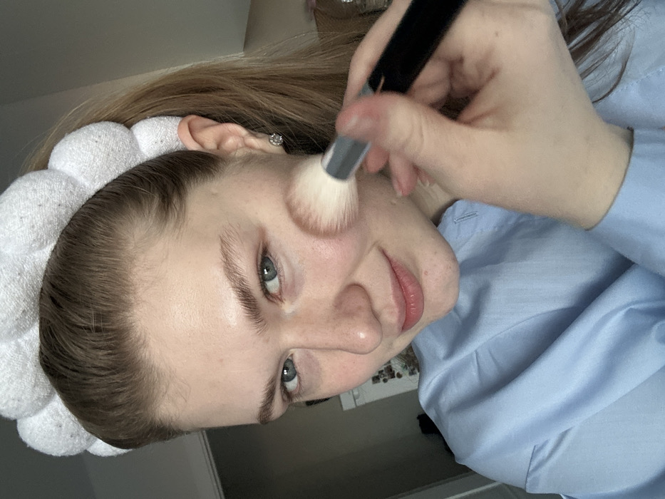 A fair-skinned woman with light brown hair and a white headband applies makeup with a brush to her cheek. She wears a light blue button-down shirt. Her face is turned slightly towards the camera with a slight smile. A silver earring is visible in her left ear. A white wall is visible behind the subject.